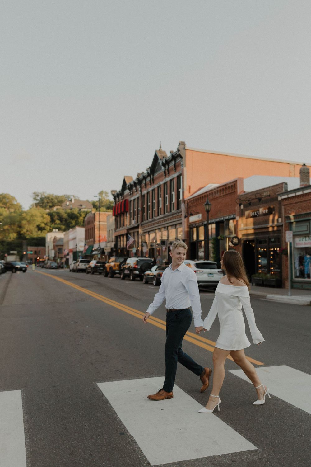 A couple walks down a charming small town street lined with historic brick buildings and parked cars.