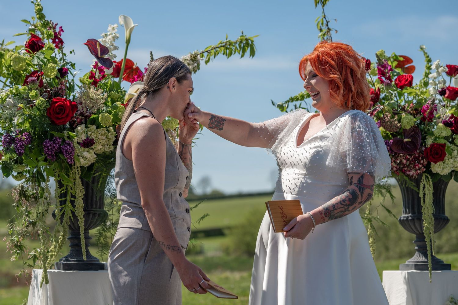 Brunette bride in tailored suit kisses the hand of a red haired bride in a white dress, standing in front of an open field and flowers