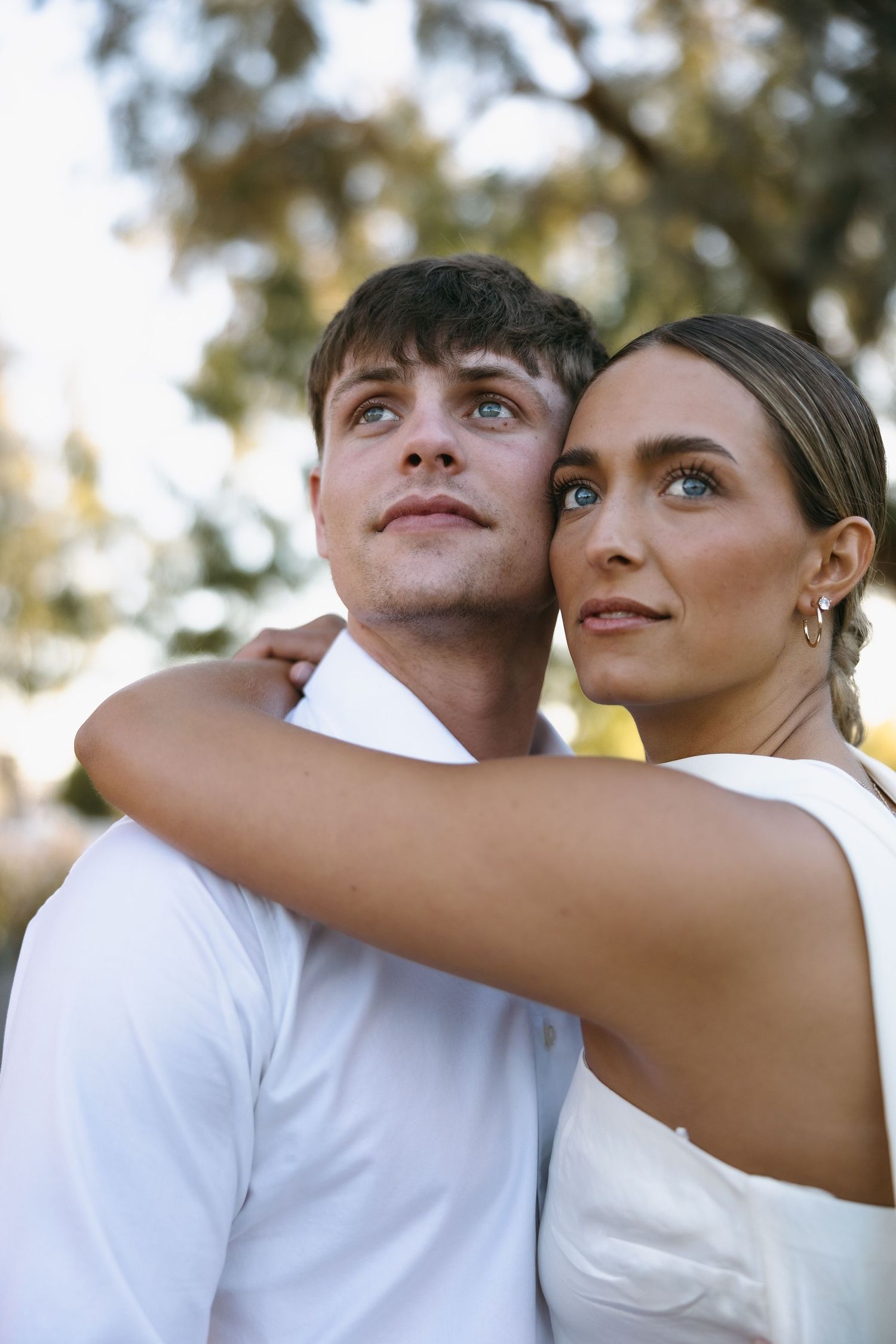 A couple in white outfits embracing while looking up at the sunset through trees.