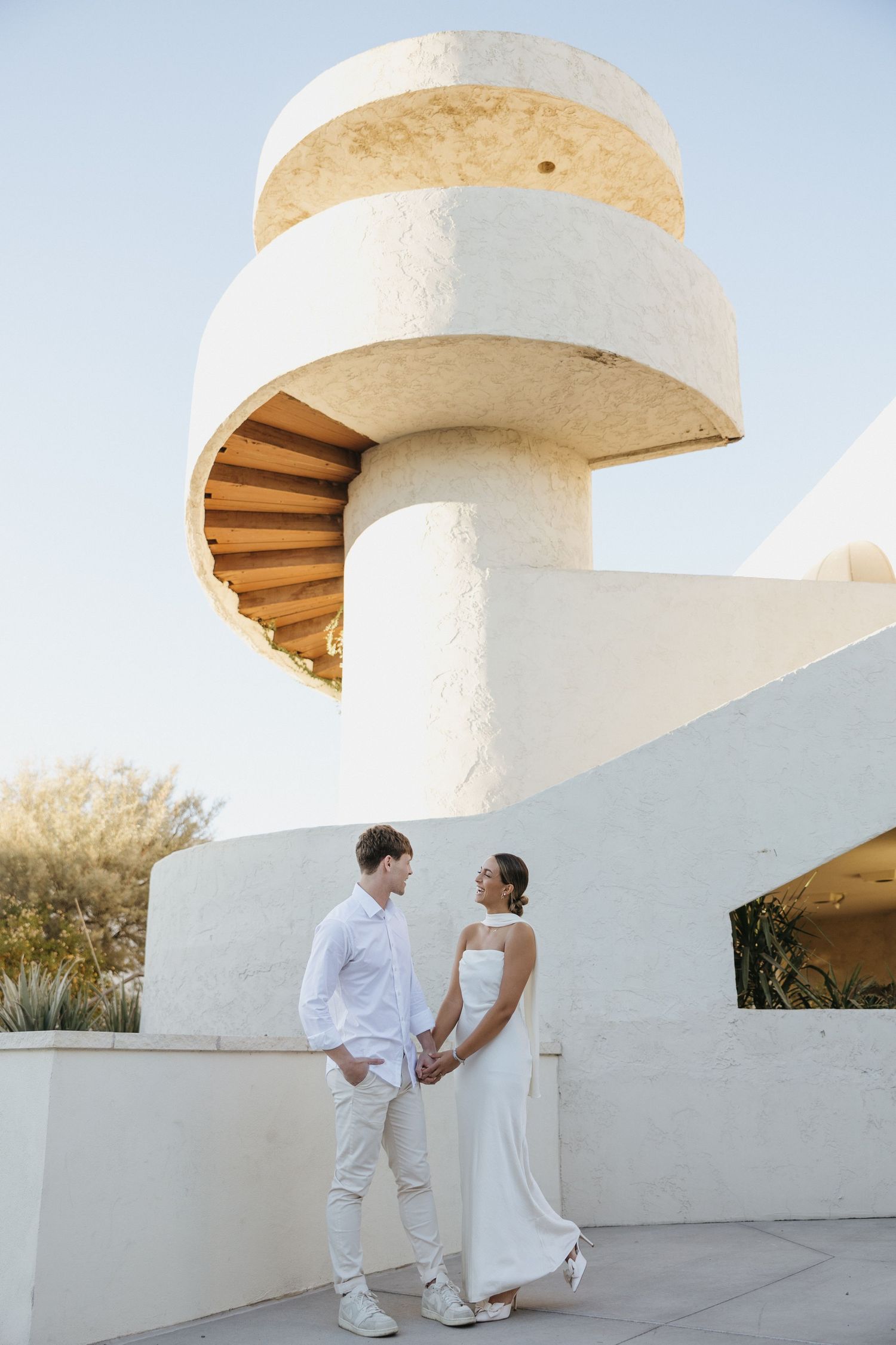 Elegant couple in white attire pose by modern spiral staircase architecture.