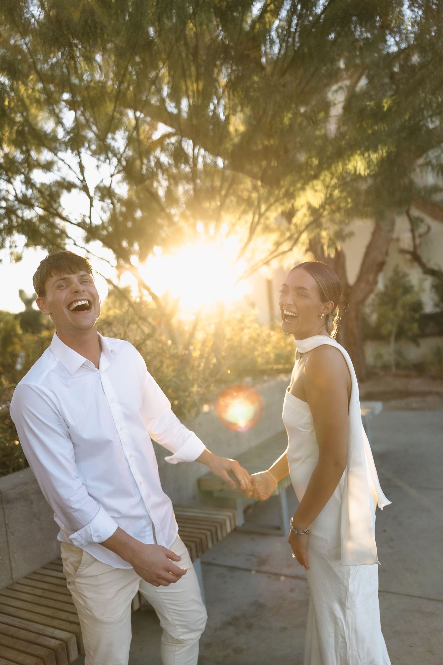 Two people in white clothing laughing together as sunlight streams through trees behind them.