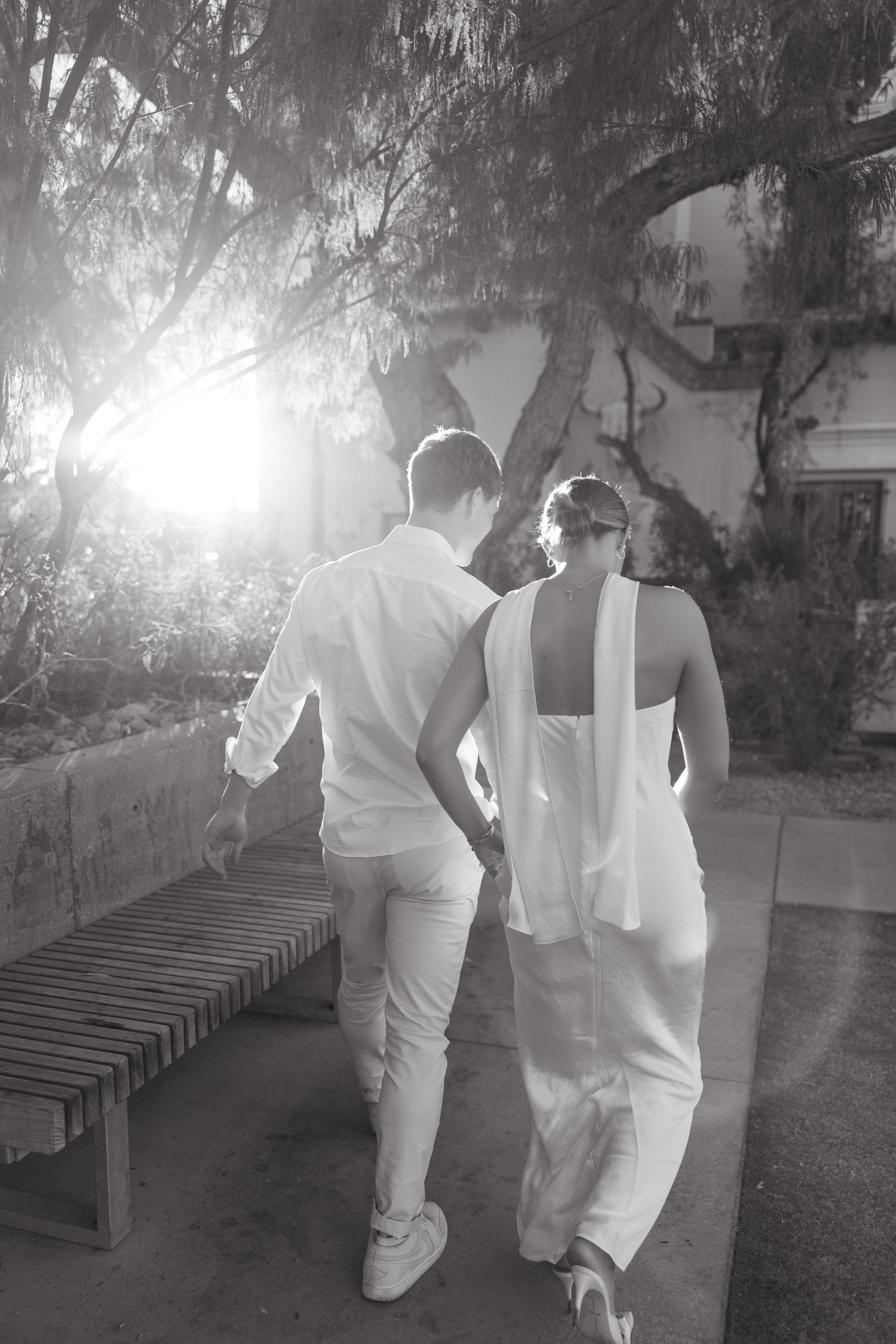 A couple in white attire walking together along a garden path at sunset.