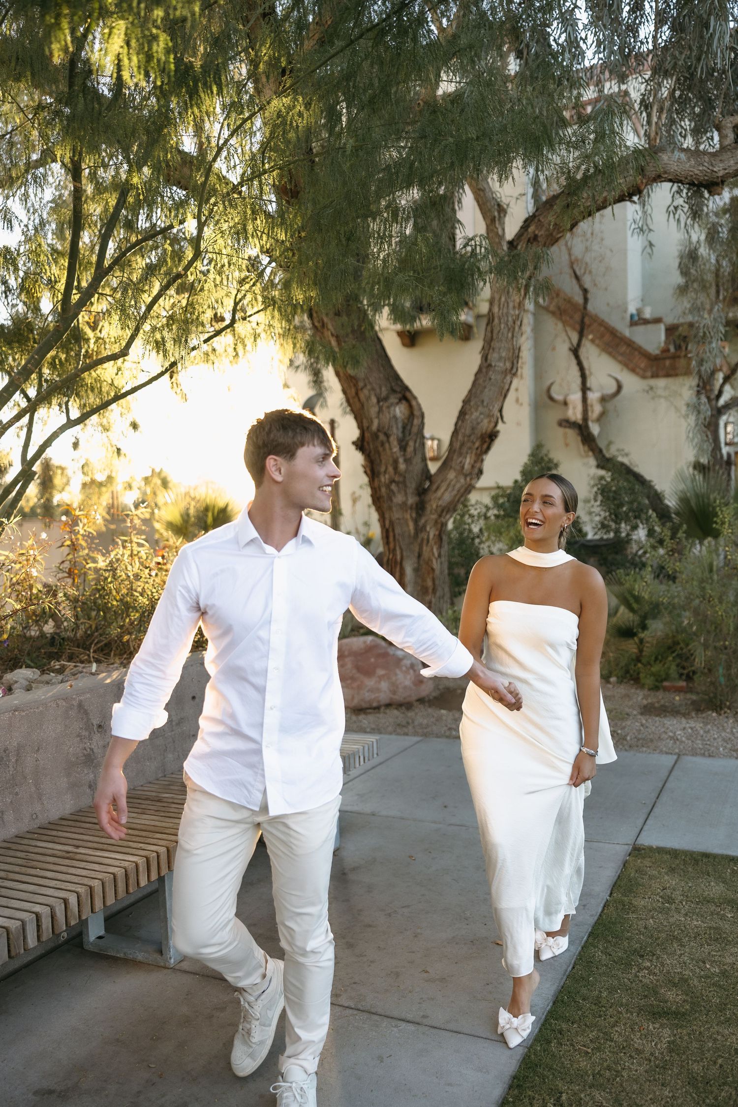 Two people in white outfits dancing playfully in a garden setting with golden sunlight.