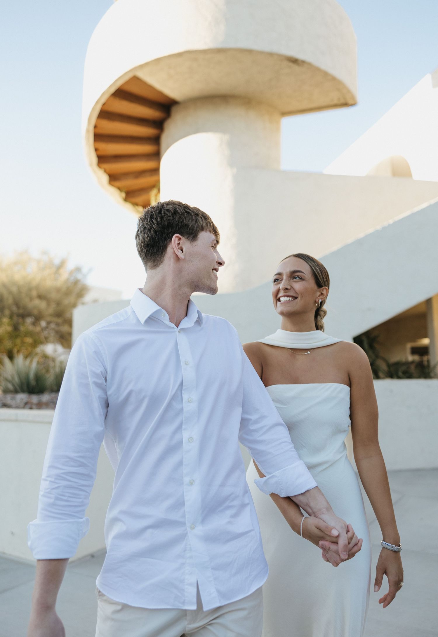 A couple in white outfits holding hands while walking near a modern architectural structure with curved details.