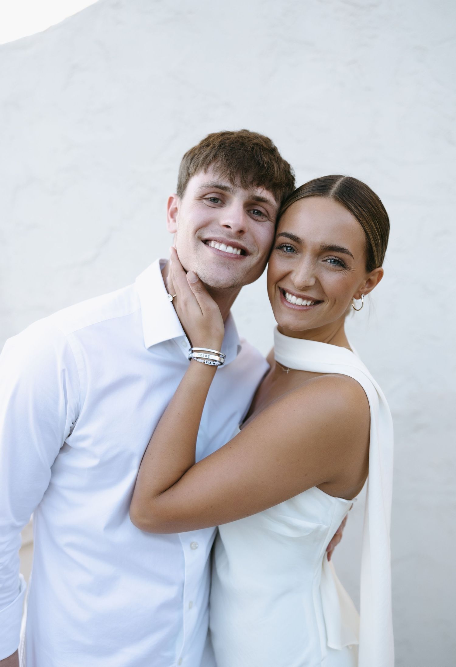 A happy couple dressed in white embracing and smiling against a light colored wall.