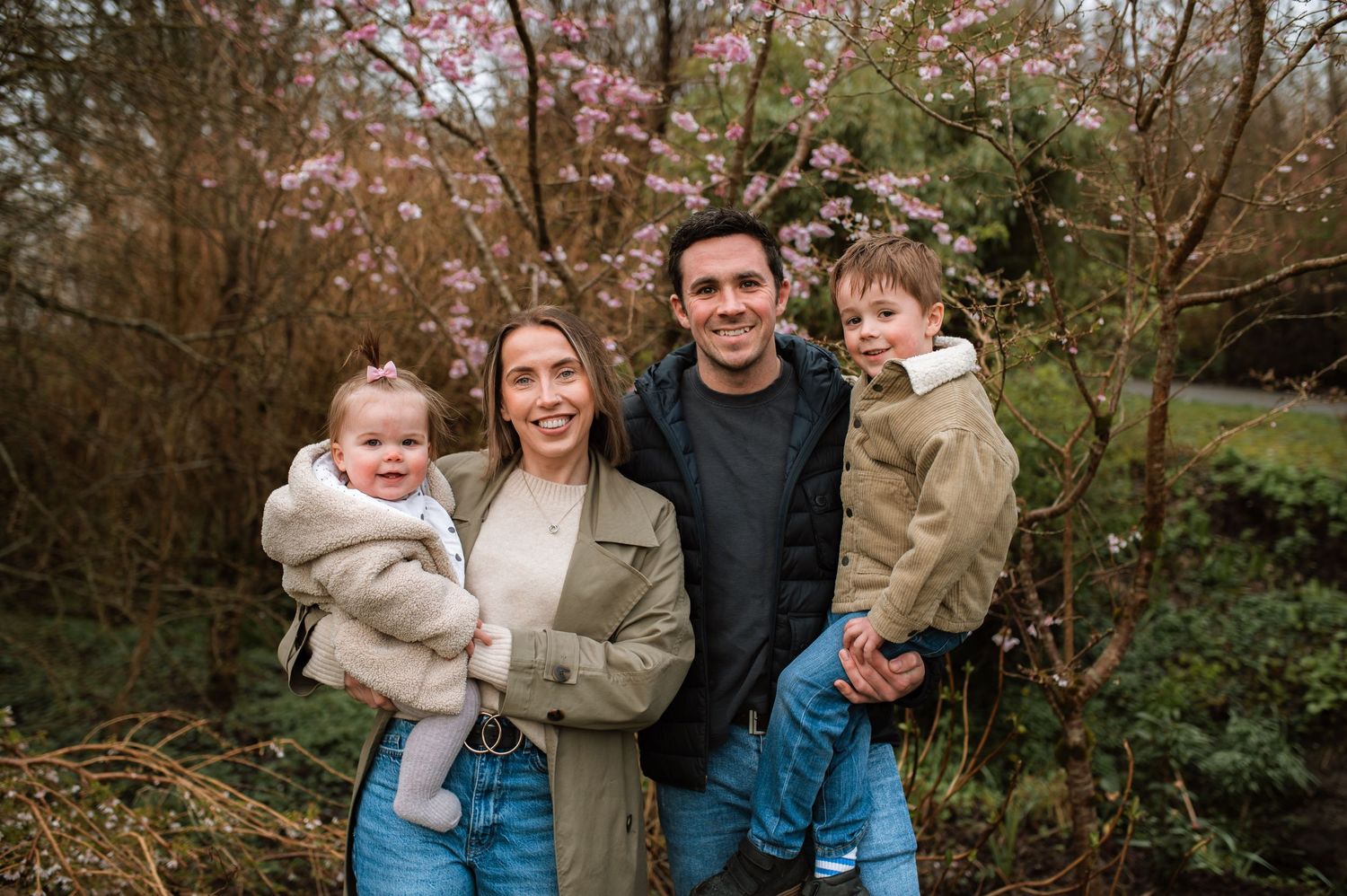 A family poses outdoors in casual clothing against a backdrop of autumn trees and foliage.