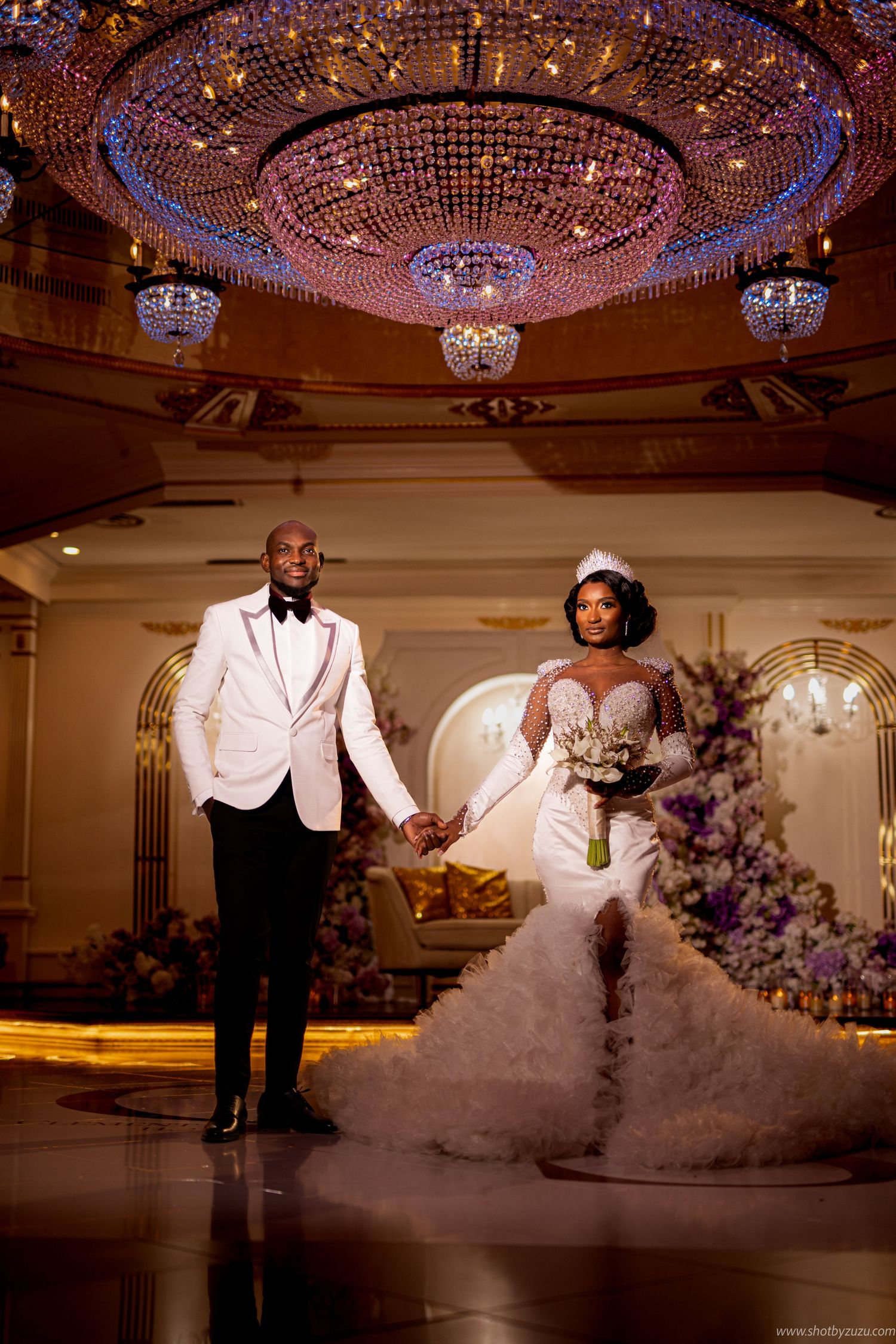 Wedding couple poses under ornate crystal chandelier in elegant ballroom setting.