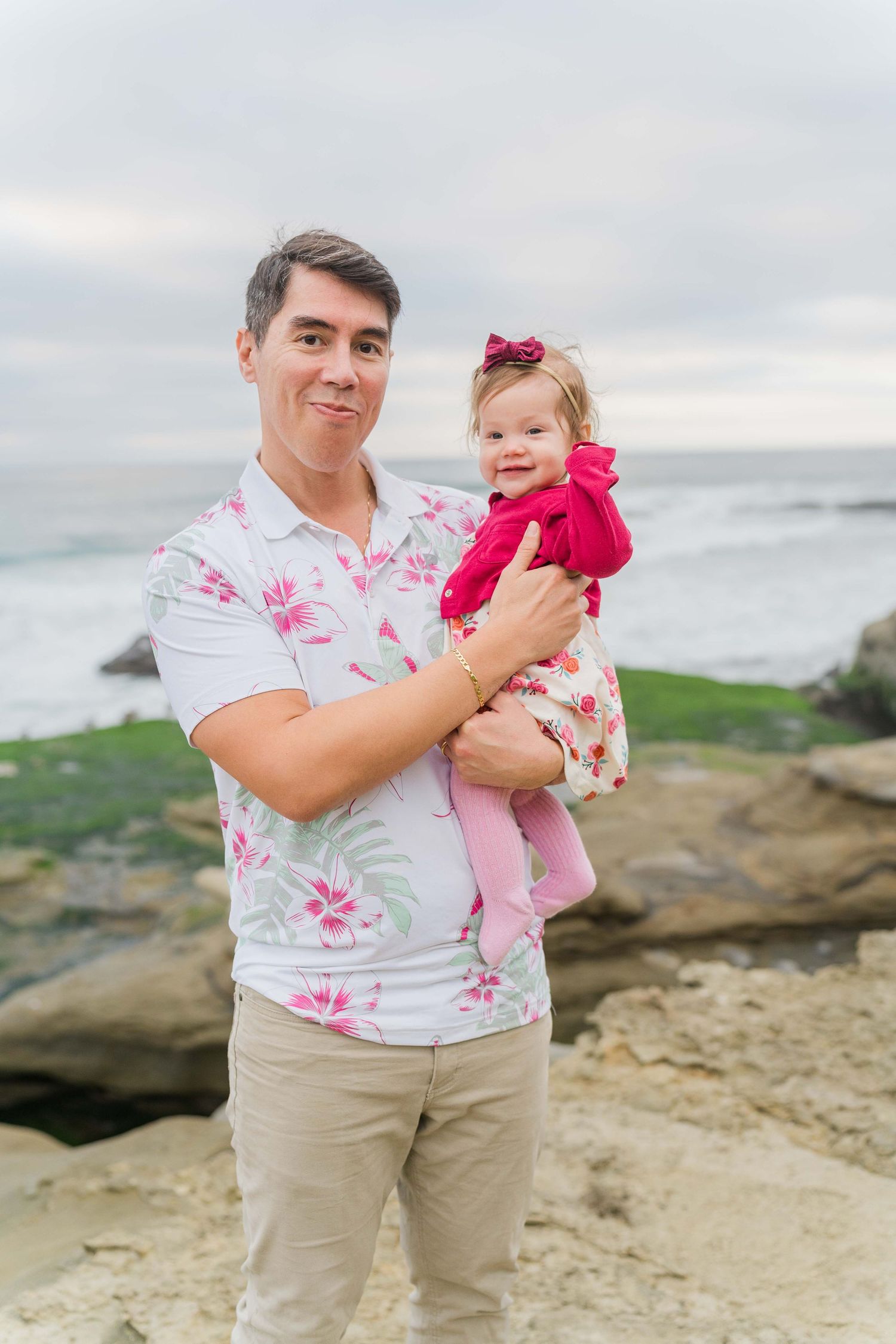 Parent holding child in pink outfit against scenic coastal background.