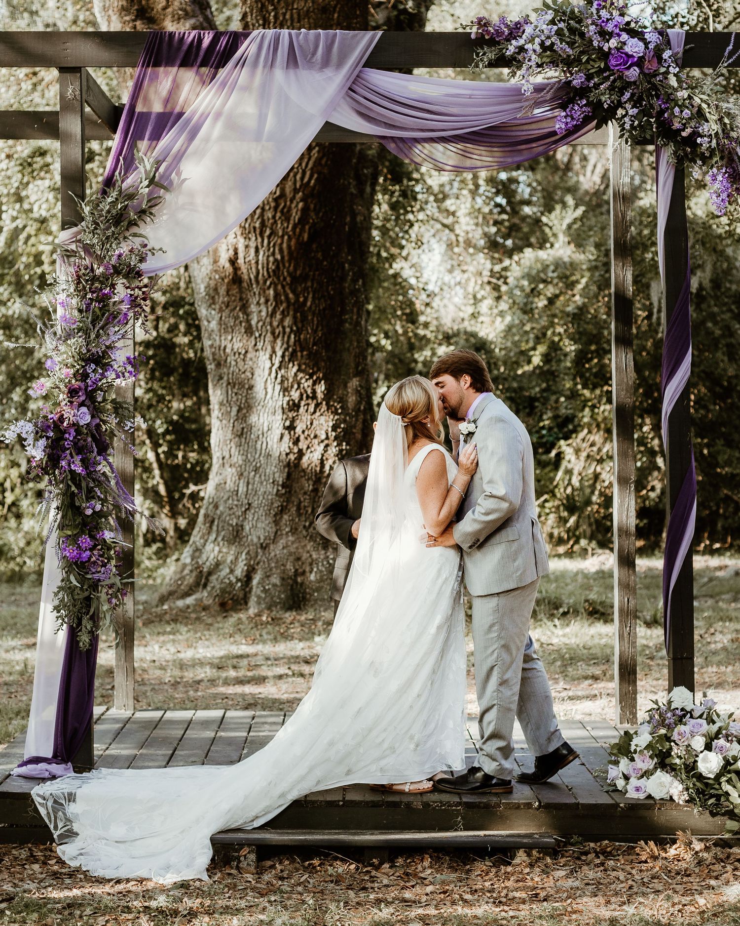 Wedding ceremony setup outdoors with purple draping and floral arrangements under mature trees.