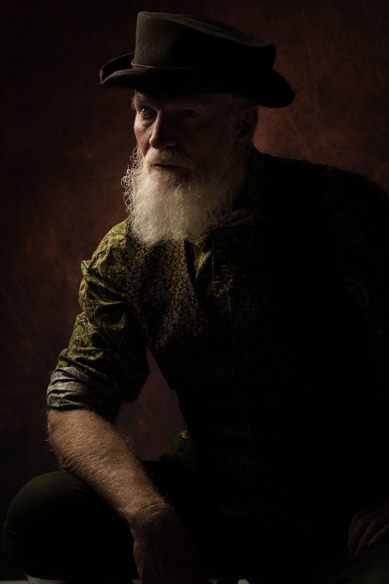 A dramatic portrait of someone with a white beard wearing a dark hat and green velvet jacket against a dark background.
