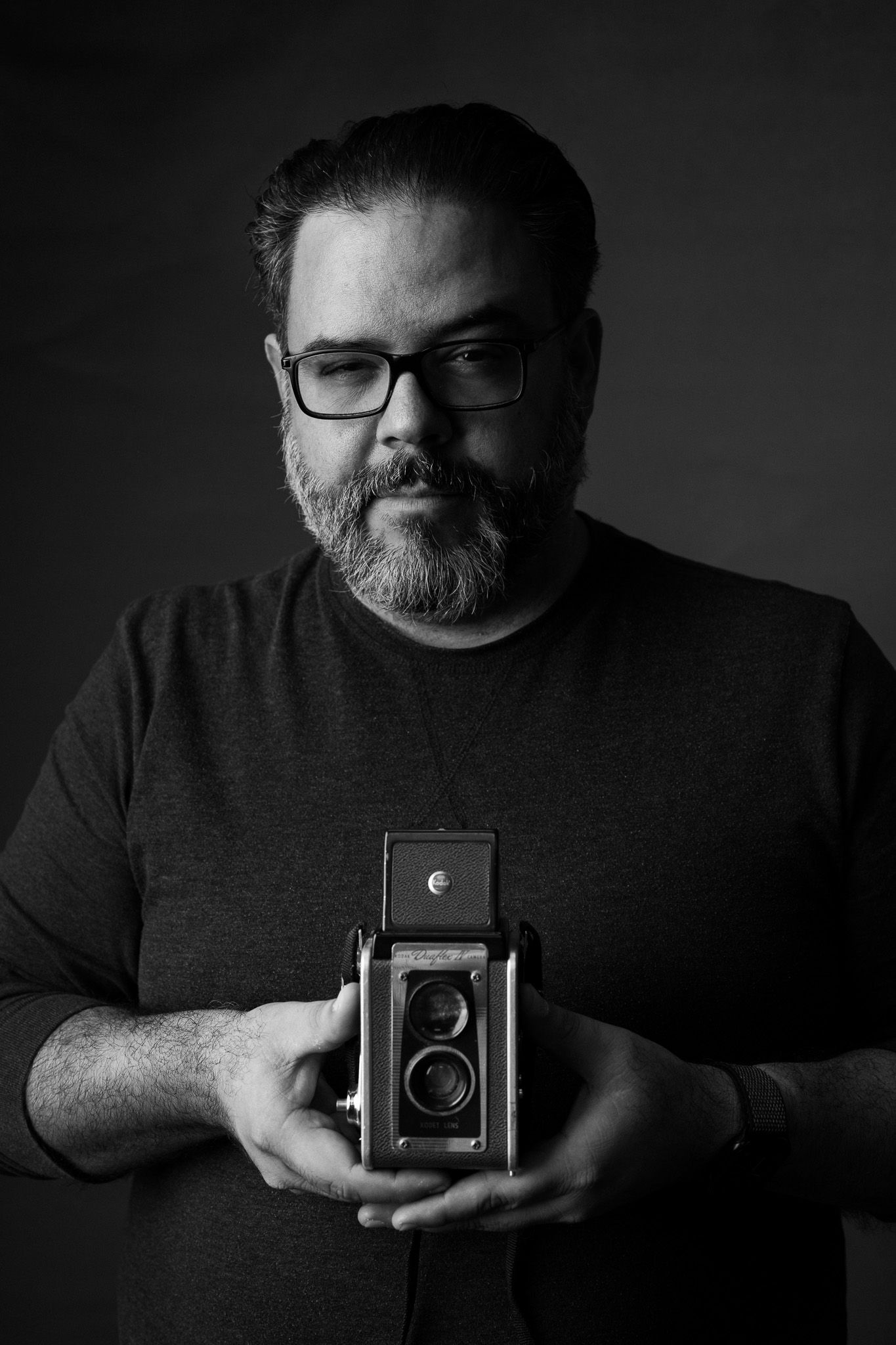 Black and white portrait of person holding vintage twin-lens reflex camera against dark background.