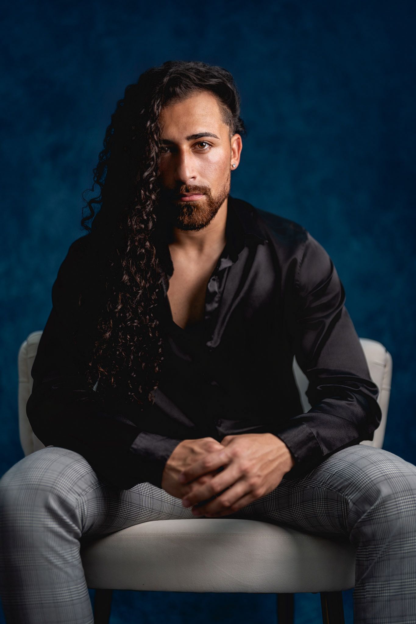 Portrait of person with long dark hair in black shirt seated in gray chair against blue backdrop.