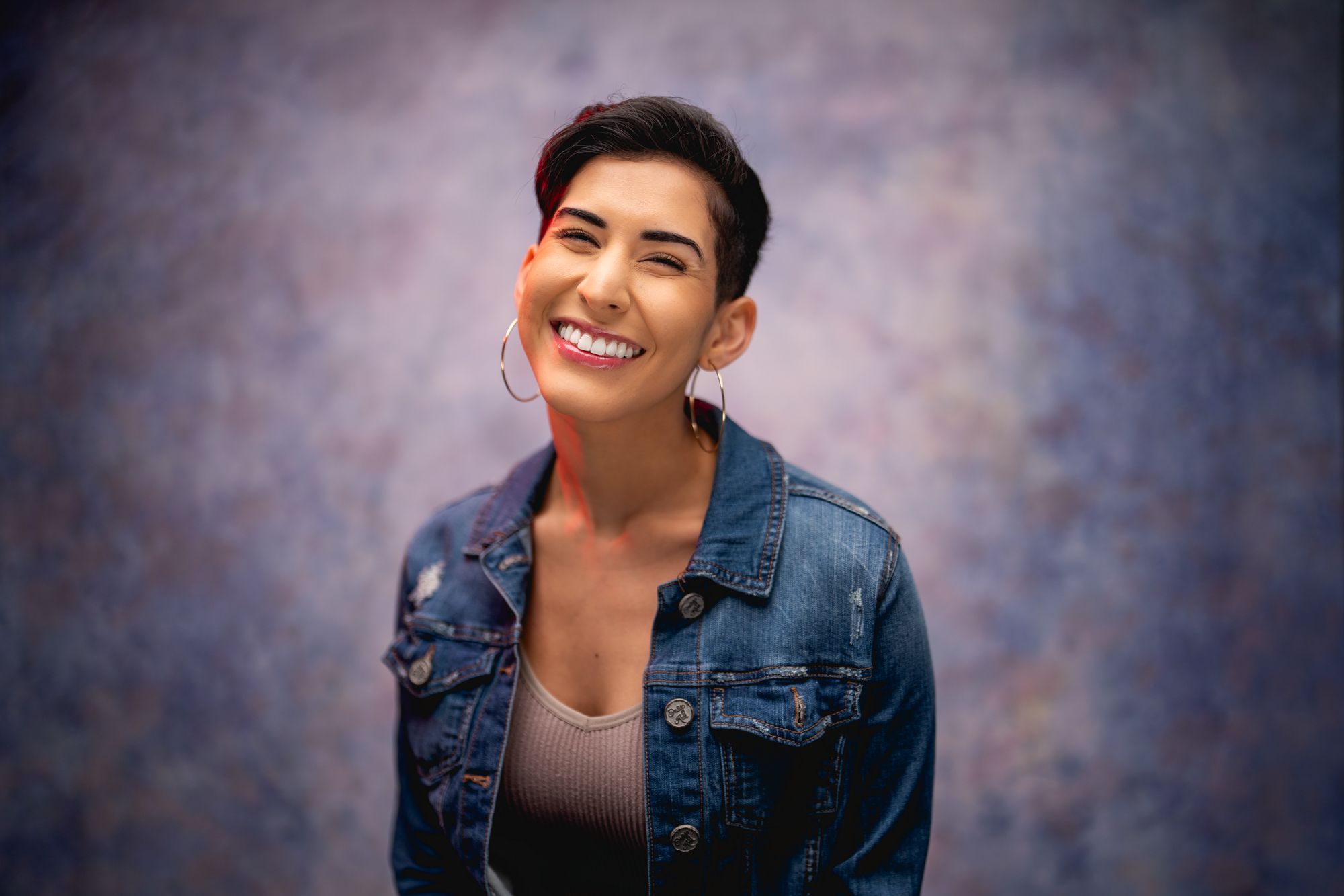 Portrait of person in denim jacket with short dark hair against mottled purple backdrop.