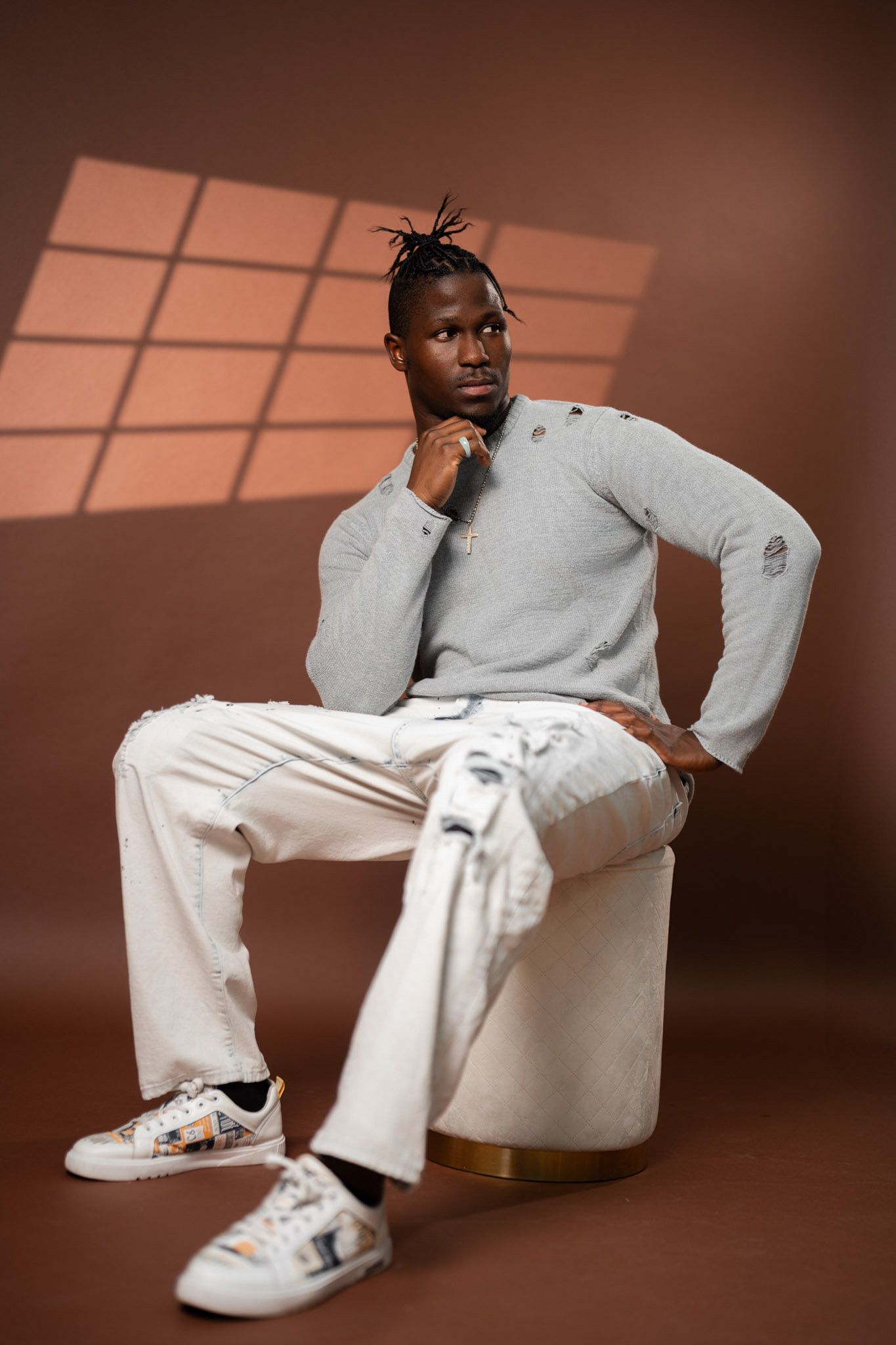 Young man in casual attire posing thoughtfully on a white ottoman against a brown backdrop with window shadows.