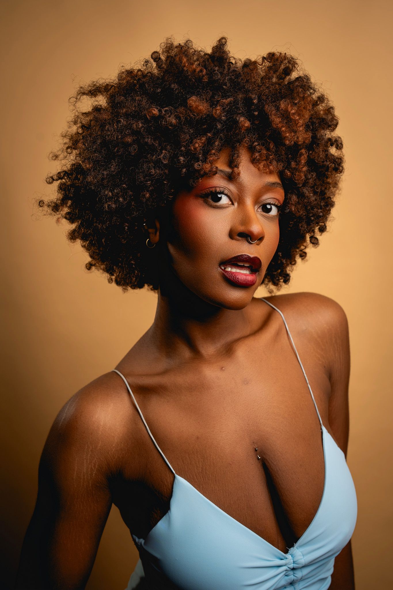 Studio portrait against warm brown background featuring beautiful natural curly afro hairstyle and light blue camisole top.