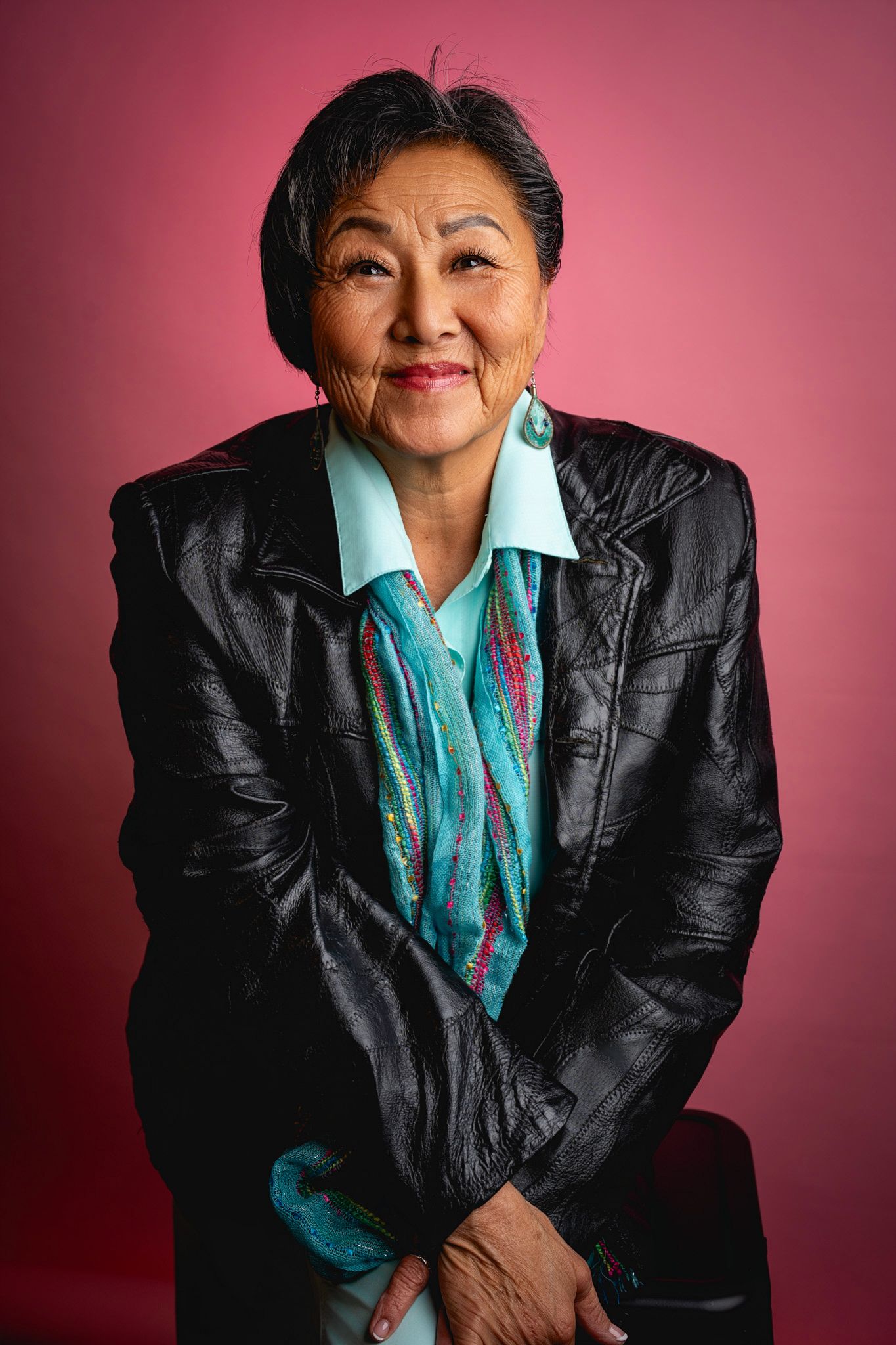 Portrait of a person in a black leather jacket and turquoise blouse against a pink background.