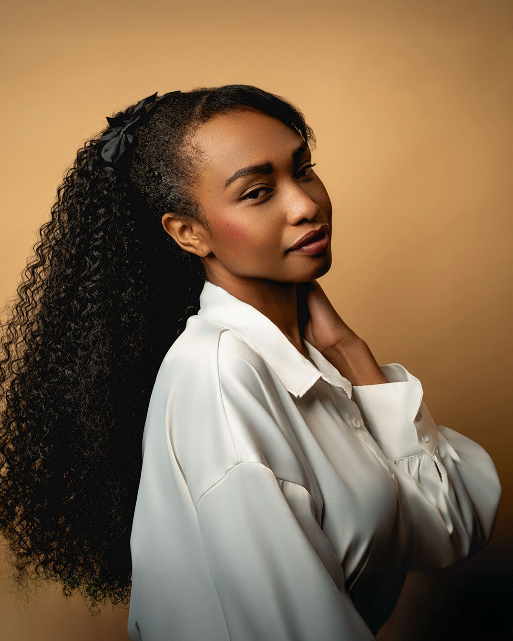 Professional studio portrait of a figure with natural curly hair wearing a white button-down shirt against a warm brown backdrop.
