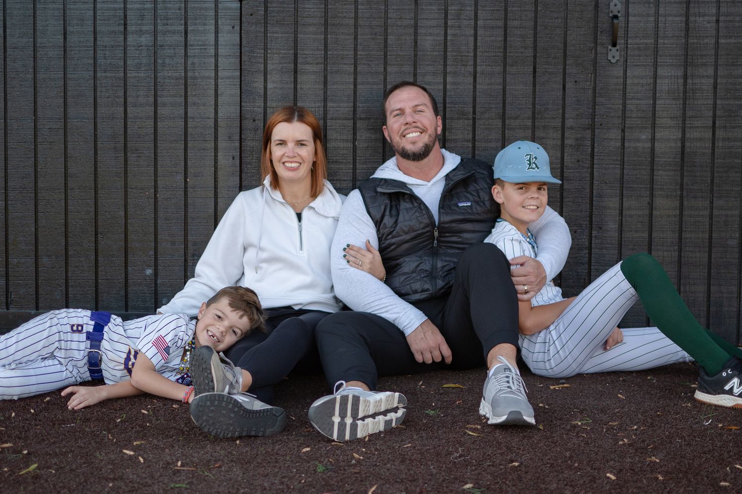 A family group sits casually together against a dark wall in baseball-style outfits and casual wear.