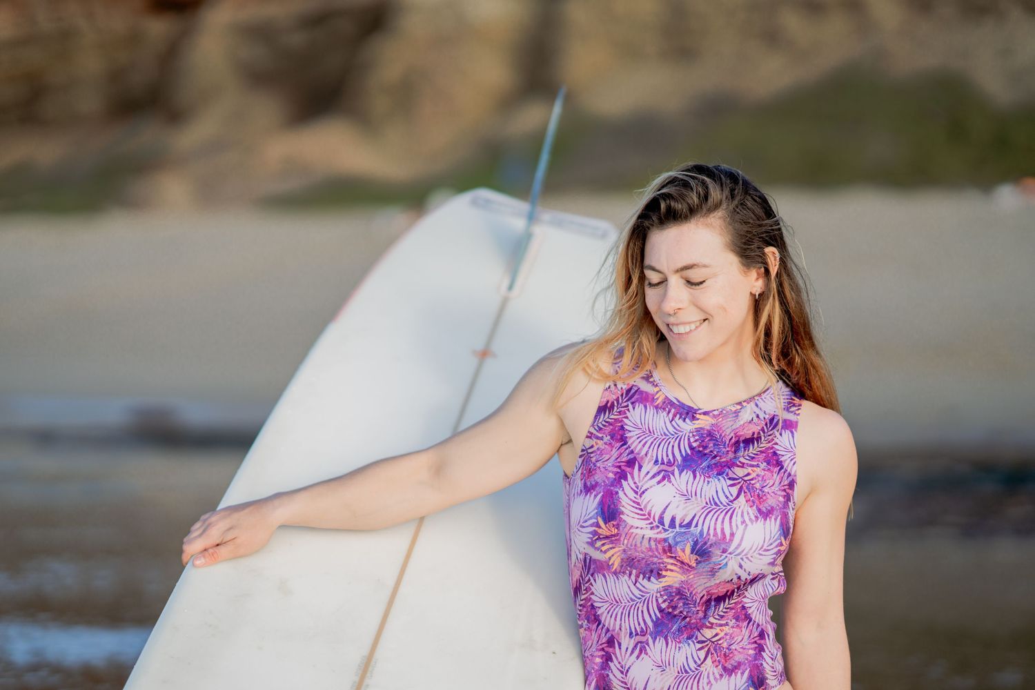 Blonde surfer girl in a one piece purple swimsuit carrying a white surfboard on her hip and smiling in Sunset Cliffs San Diego