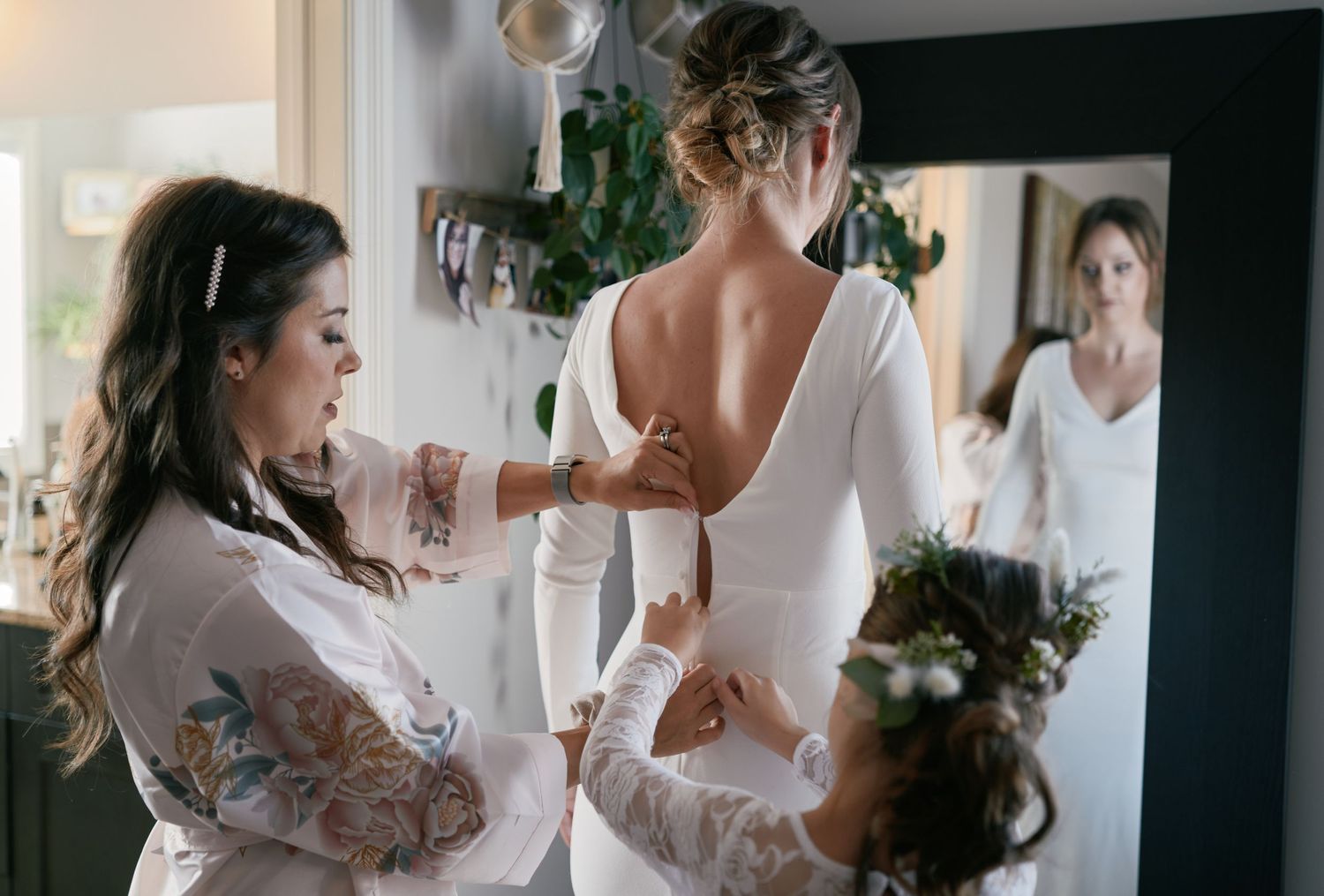 A bride gets ready as loved ones button up her elegant gown, with her reflection in the mirror capturing the anticipation of the moment.
