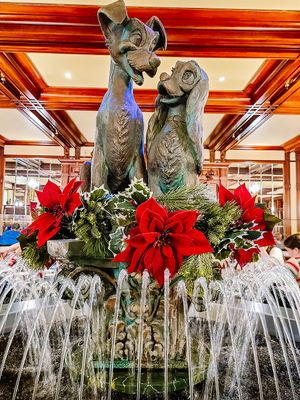 Iconic Disney Lady and The Tramp statue decorated with festive red poinsettias and holiday greenery at water fountain display.