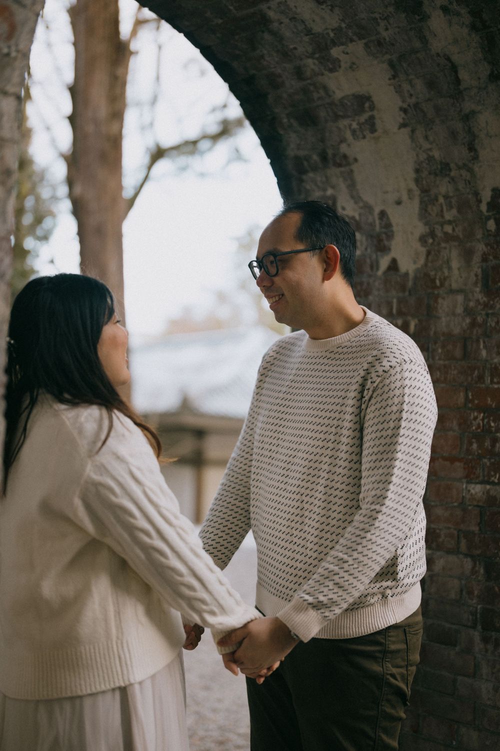 Asian couple hold hands and share a tender gaze while standing beneath a weathered brick stone arch.