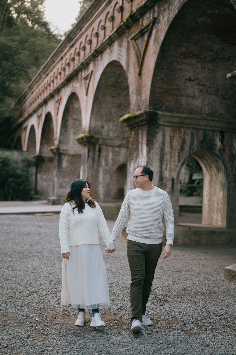 Couple holding hands walking in front of historic brick arched viaduct wearing coordinating white and neutral outfits.