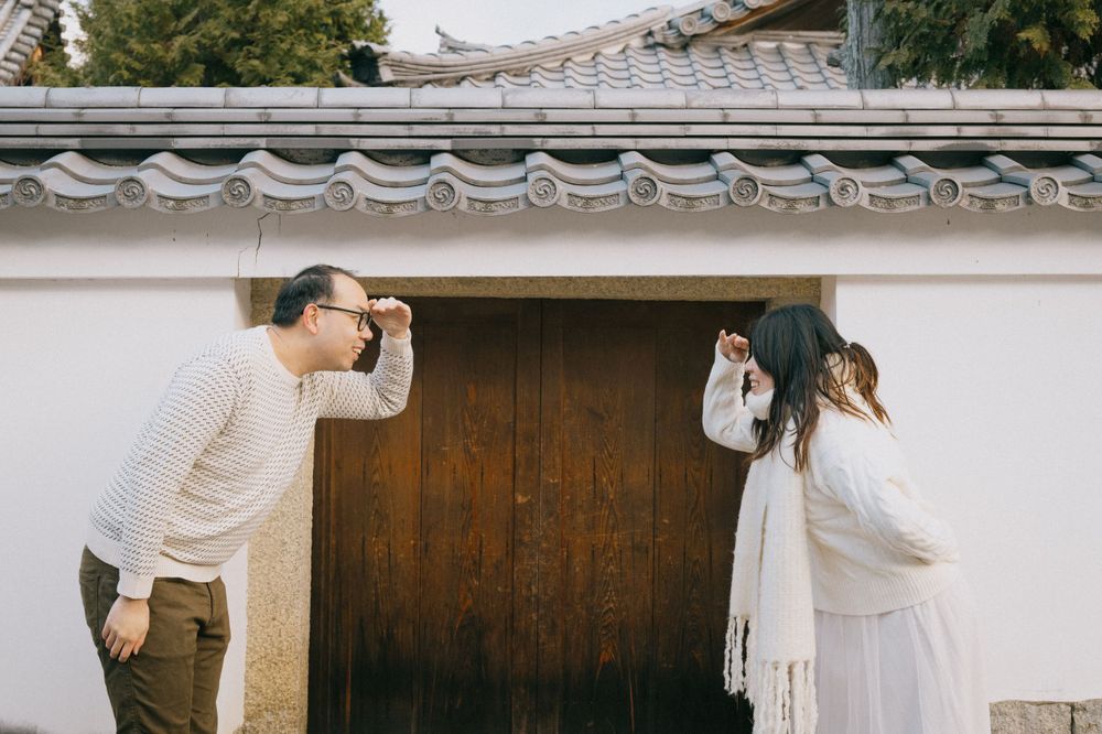 Couple playfully posing in front of wooden door beside white Japanese wall with tiled roof.