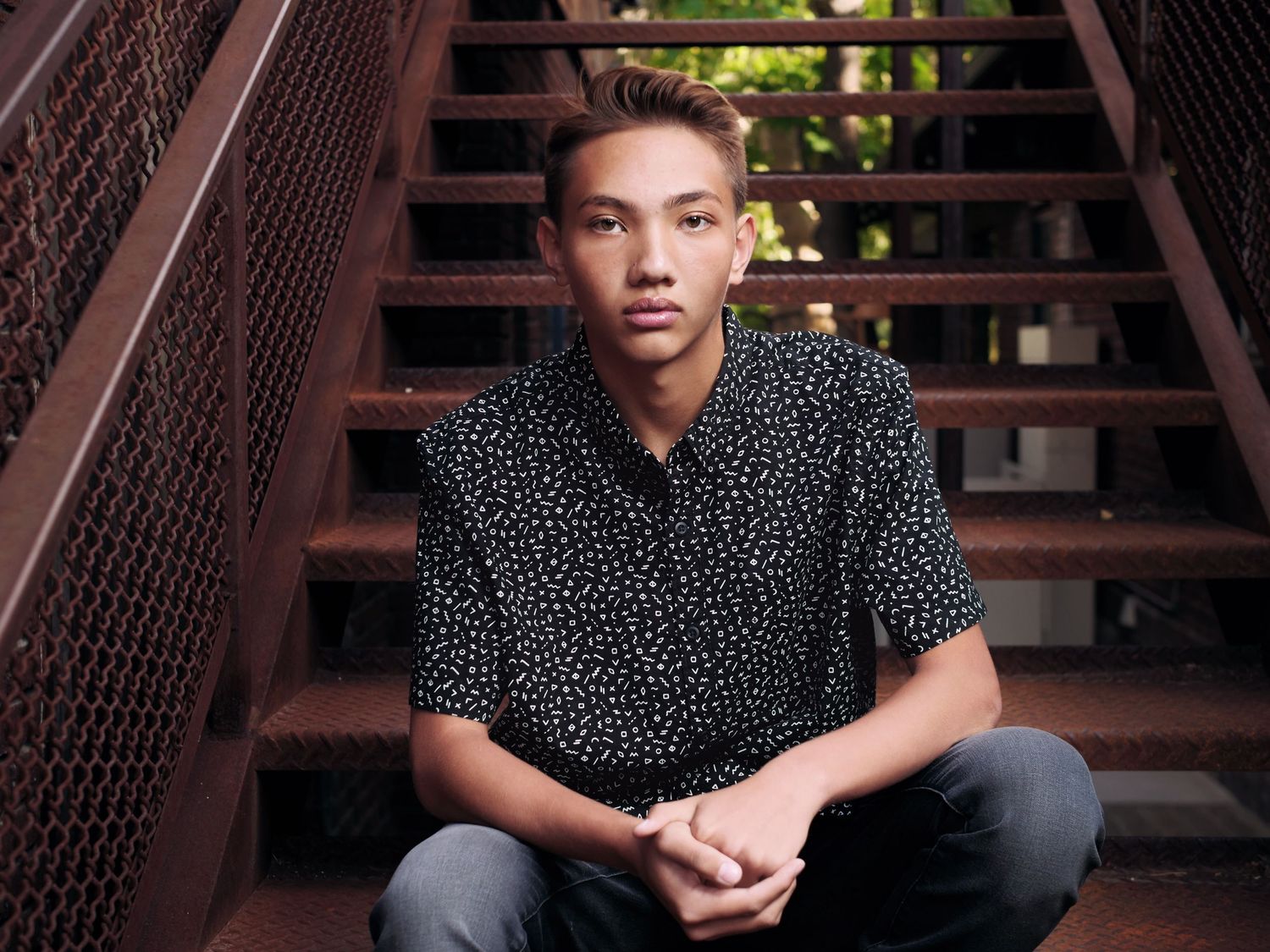 Youth wearing a black bold patterned shirt sits on rustic metal stairs with hands clasped in casual portrait pose.