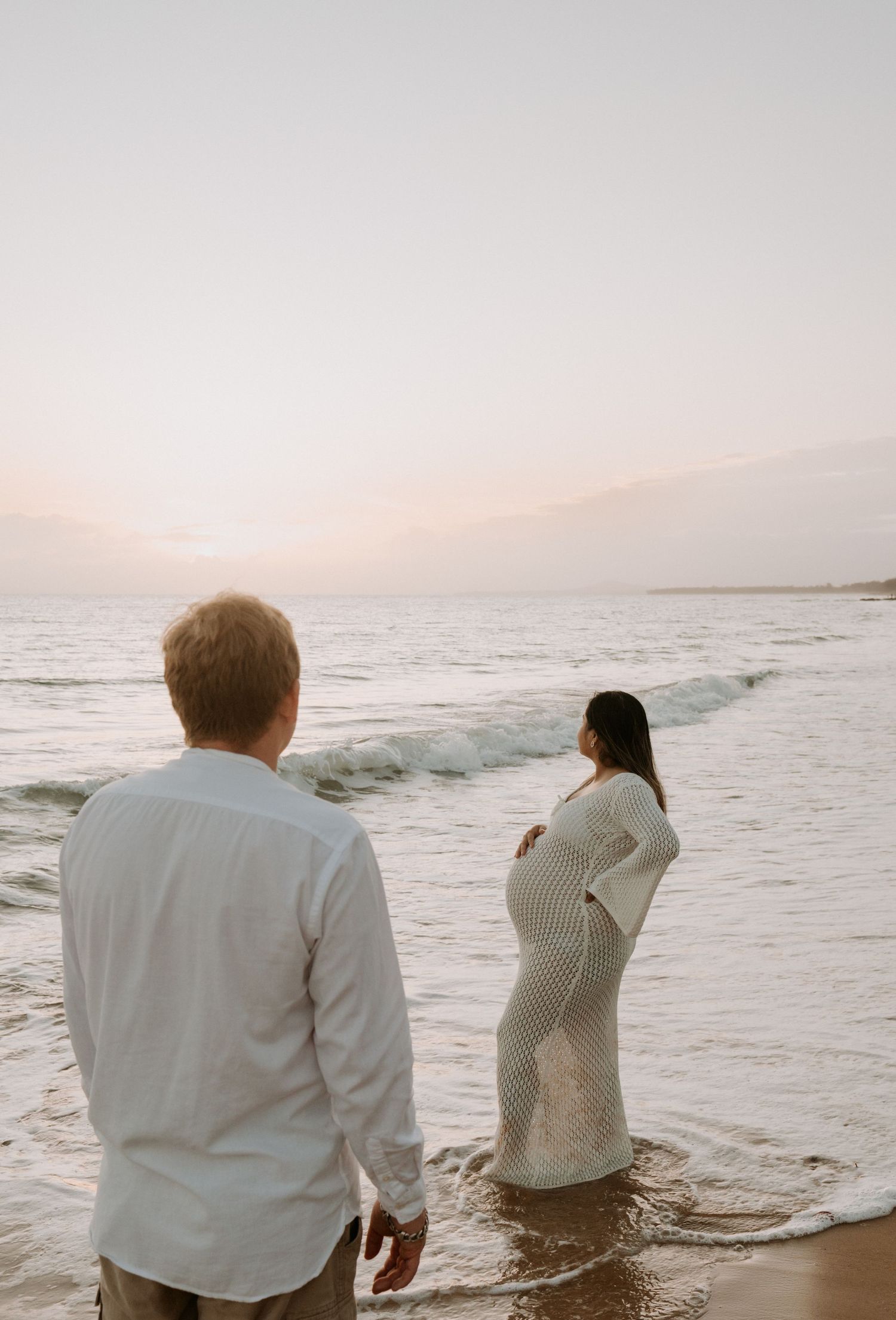 Couple stands by ocean waves at sunset with woman in knit dress.