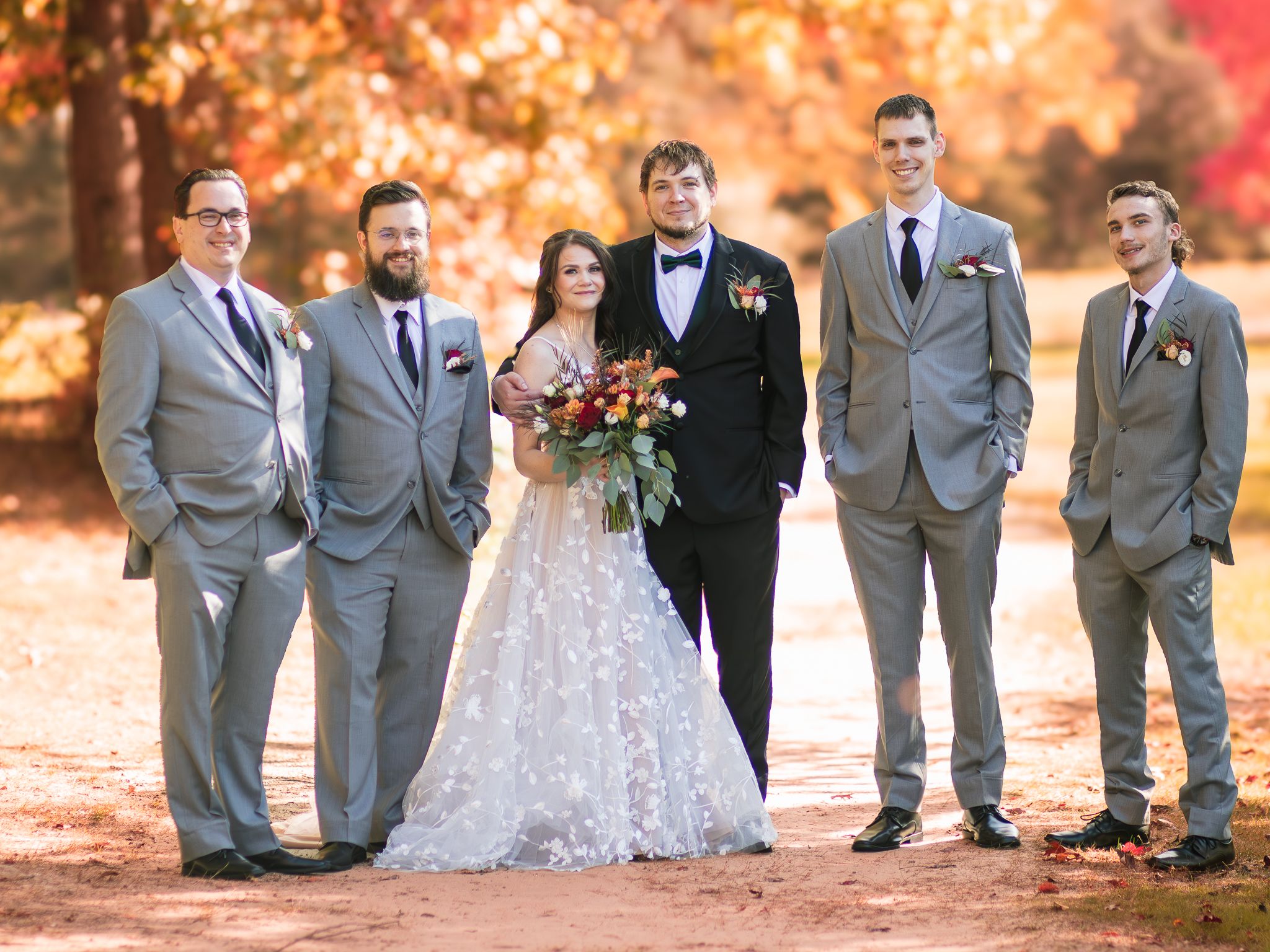 Wedding party poses outdoors on autumn path with fall foliage in warm golden light.