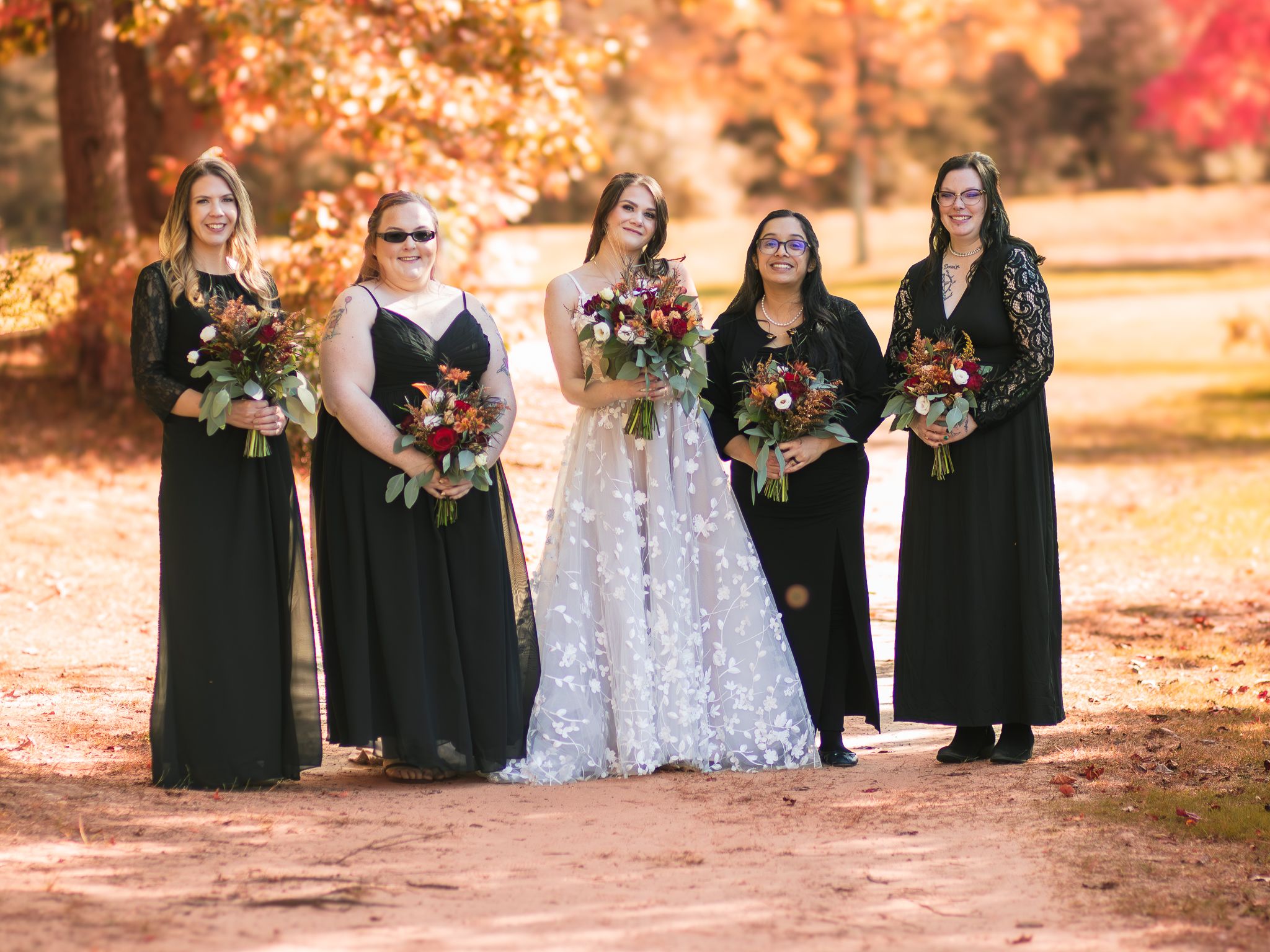 Bridal party in black dresses and white gown stand together on path with autumn trees behind them.