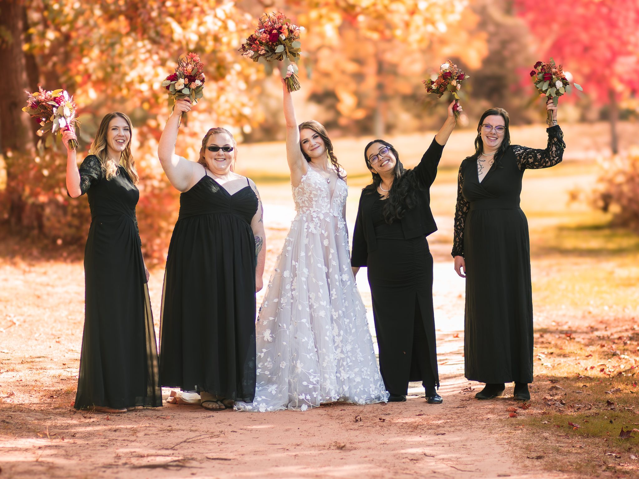 Bridesmaids in black dresses raise bouquets while celebrating with bride in white lace gown during fall photo shoot.