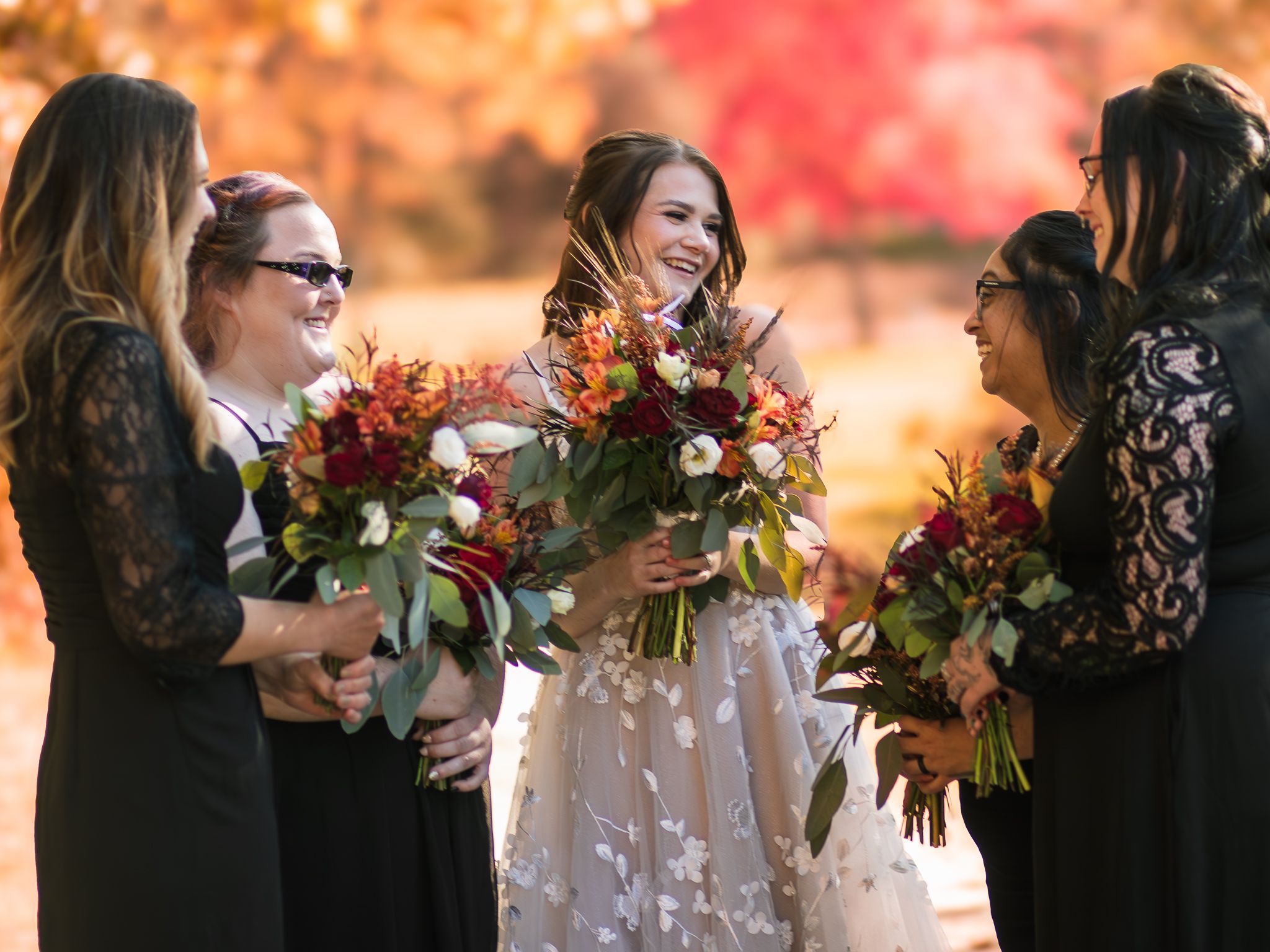 Wedding party in black dresses holding vibrant fall bouquets against autumn foliage.