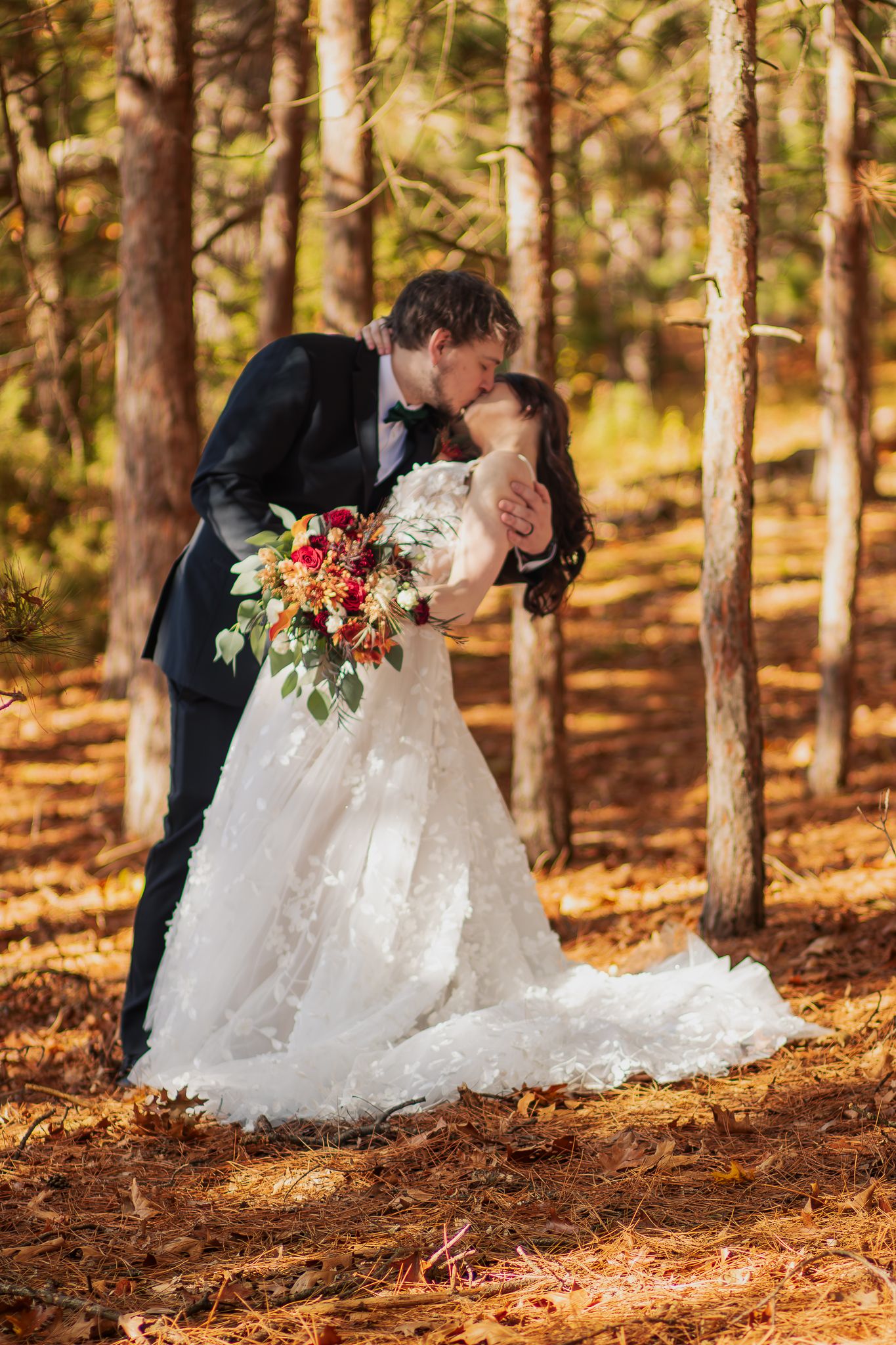 A romantic wedding couple shares a kiss in a sunlit autumn forest with pine trees and fallen leaves.