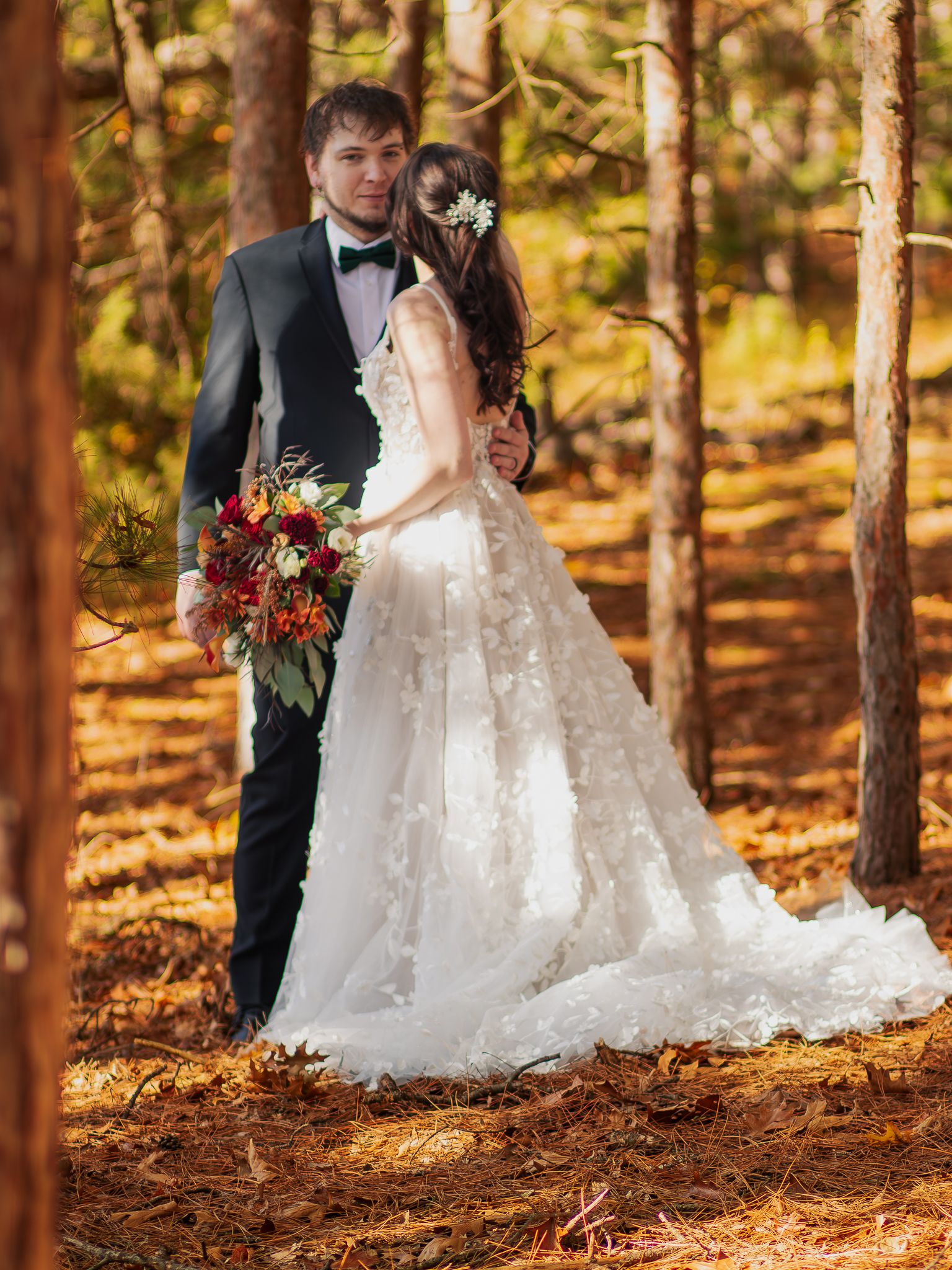 A romantic autumn wedding photograph of a couple in formal attire standing among golden-leaved trees in a forest setting.