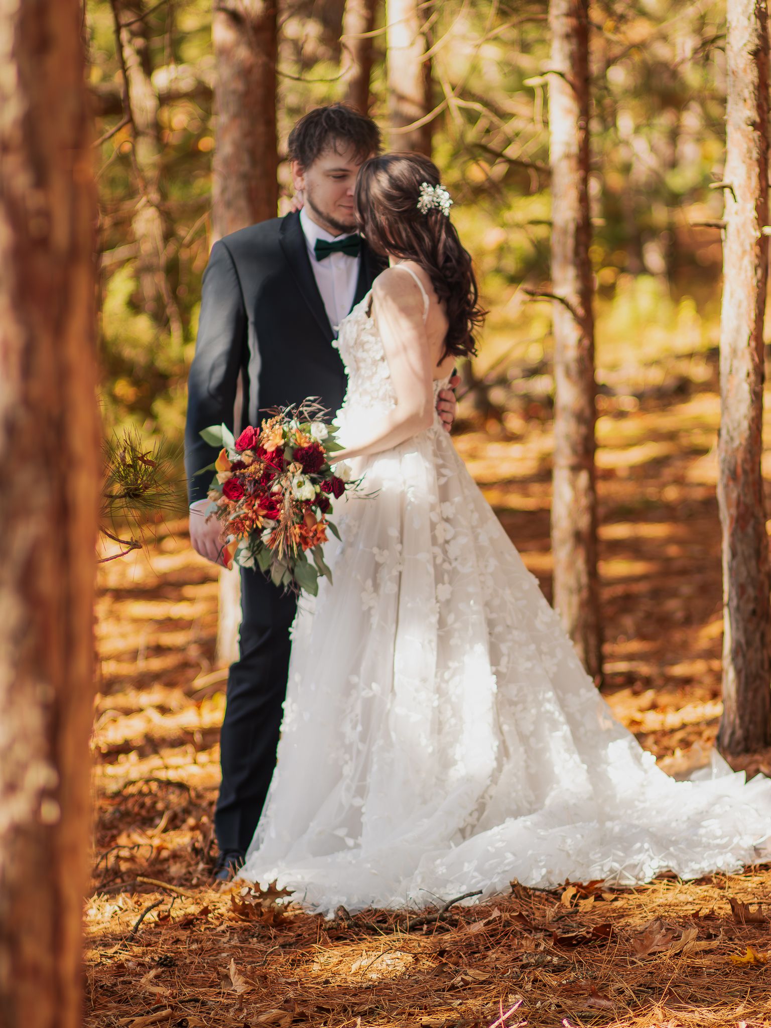 Newlywed couple share intimate moment in sunlit forest with bride holding vibrant autumn bouquet.