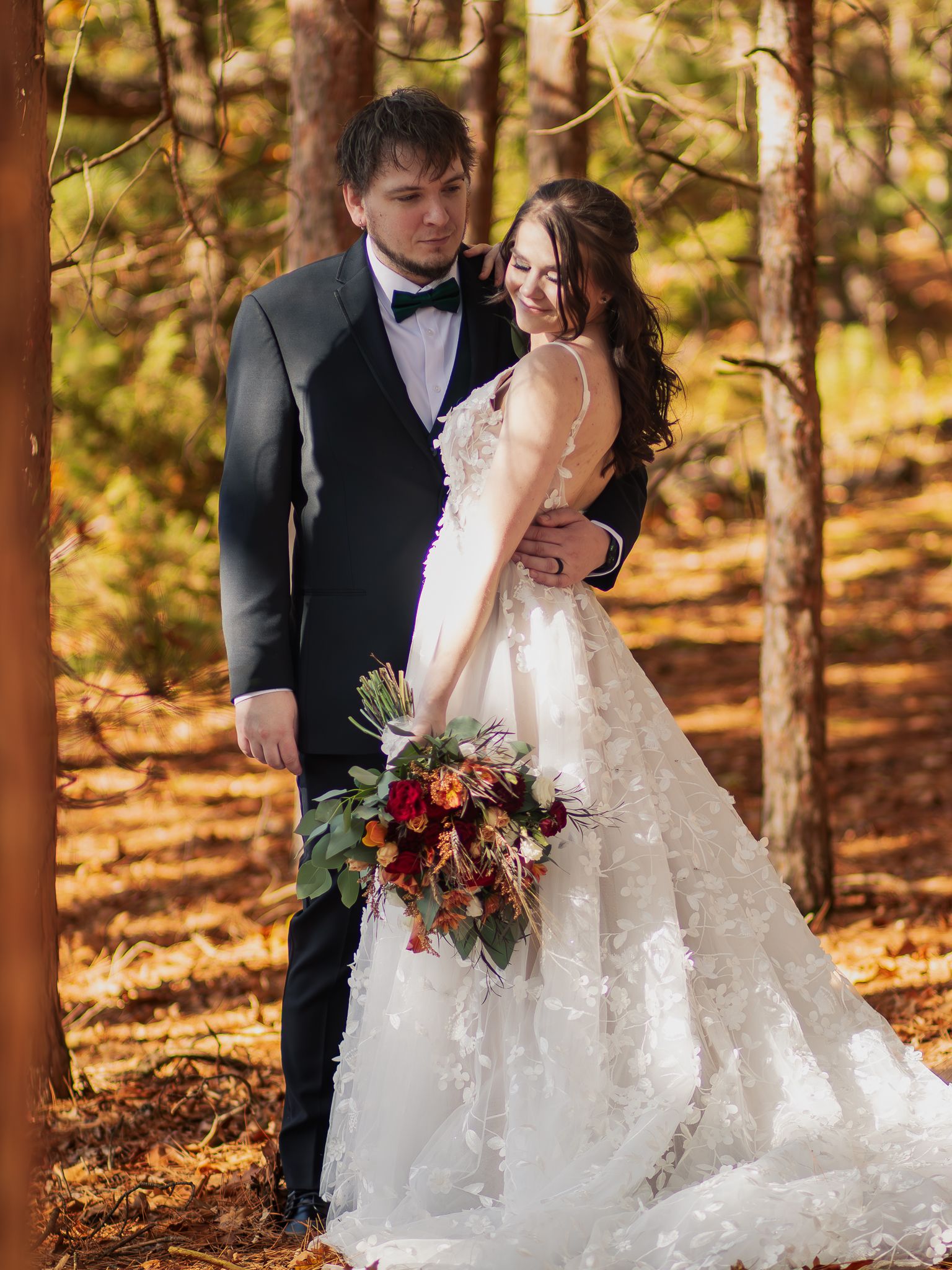 A couple poses in formal wedding attire among autumn trees with a vibrant fall bouquet of red and burgundy flowers.
