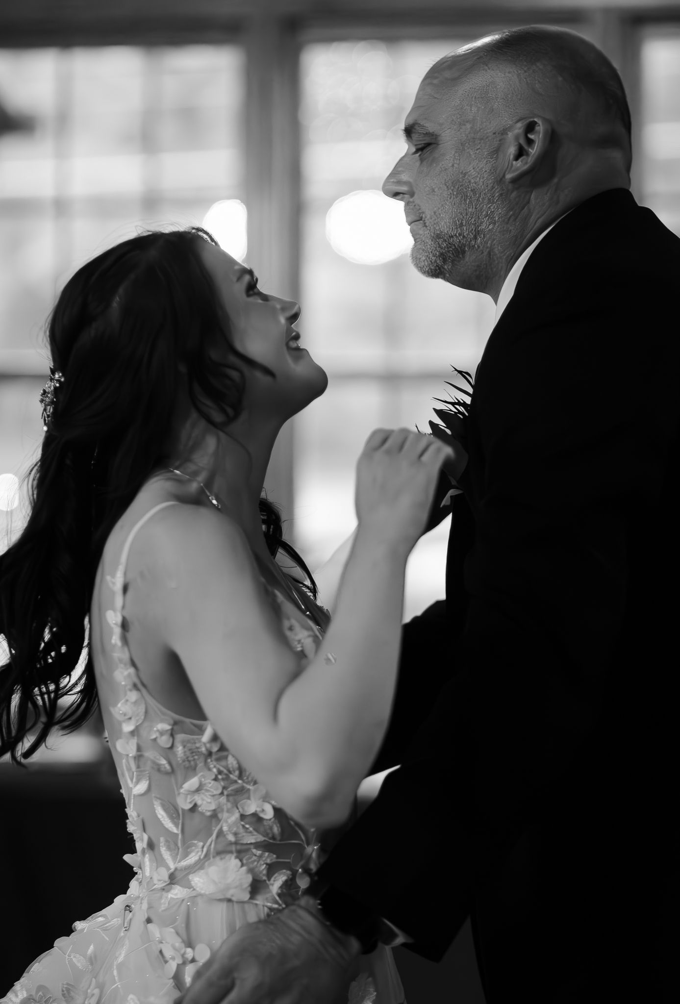 A bride and groom share an intimate moment in black and white, with the bride in a detailed lace dress touching his lapel.