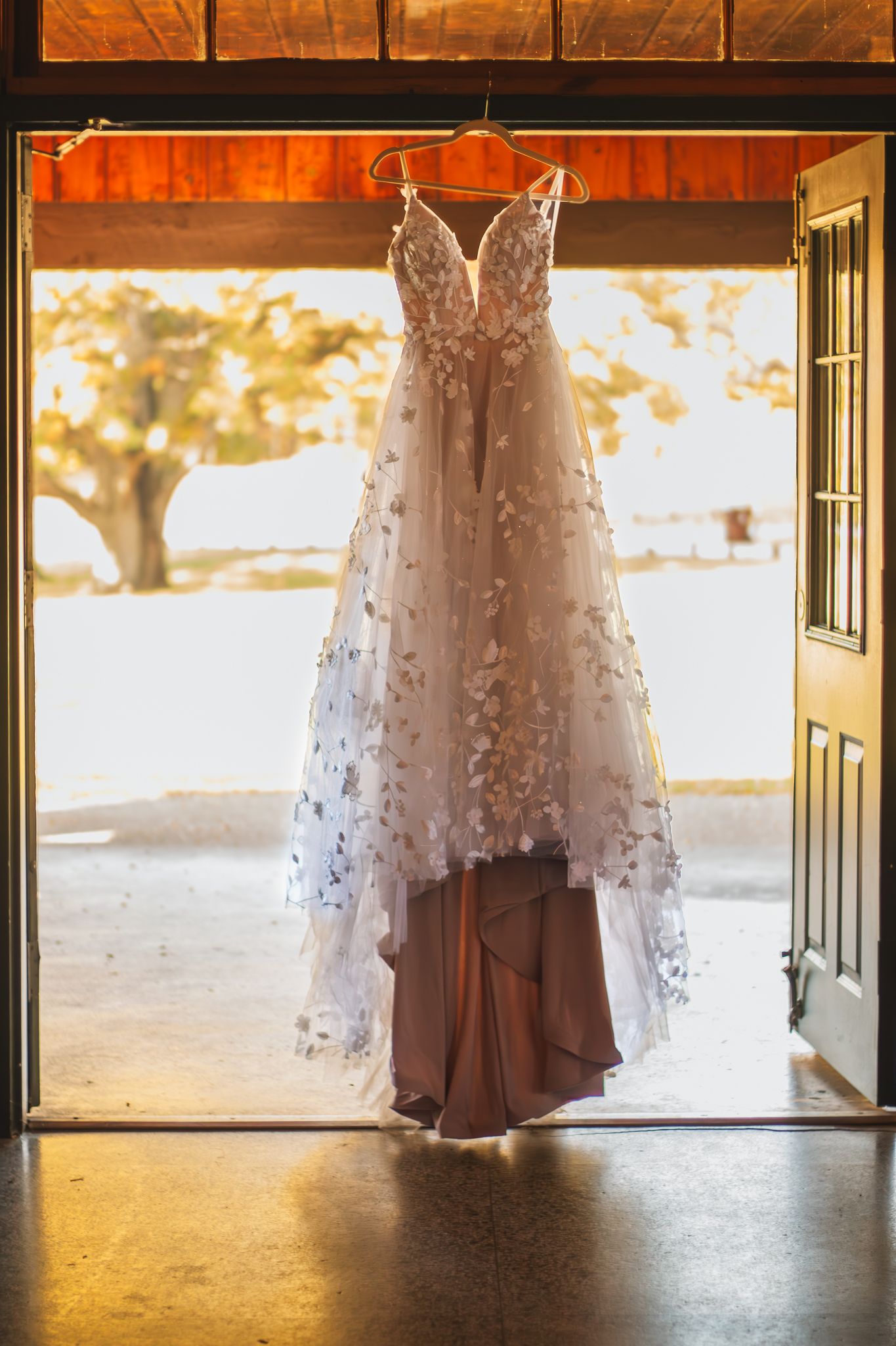 A delicate white lace wedding dress with floral details hangs in a doorway backlit by warm golden sunlight.