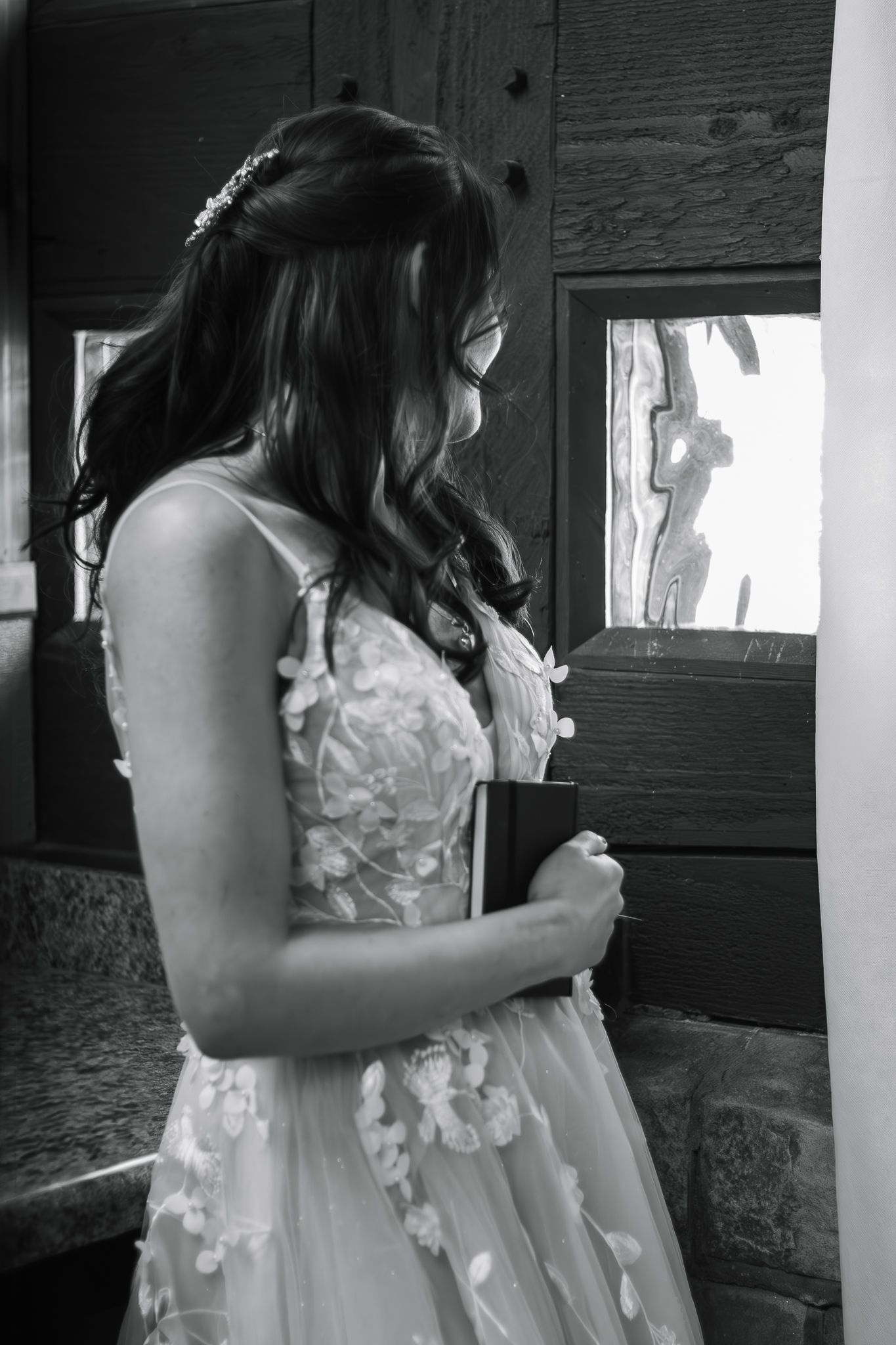 A bride in a delicate floral wedding gown stands by a window in black and white photography.