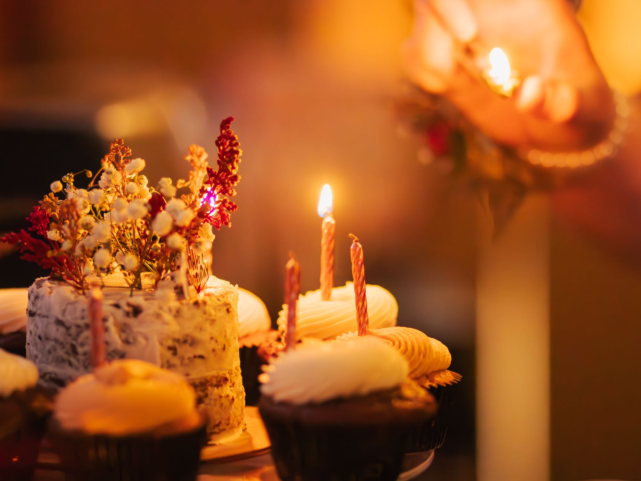 Lit birthday candles glow warmly on cupcakes decorated with colorful toppings in a dimly lit setting.