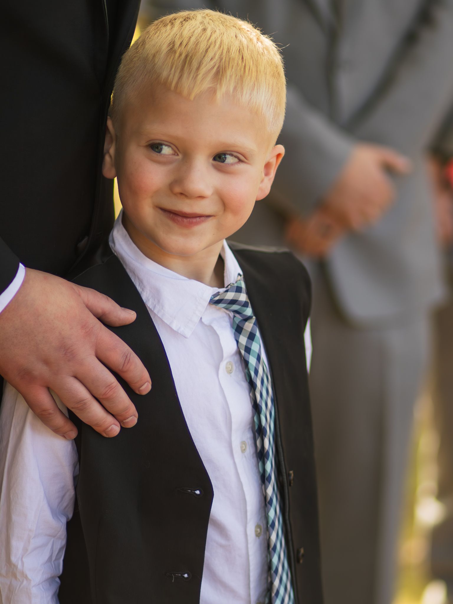 Young child dressed formally in a black vest, white shirt and plaid tie smiles at camera with a gentle hand on shoulder.