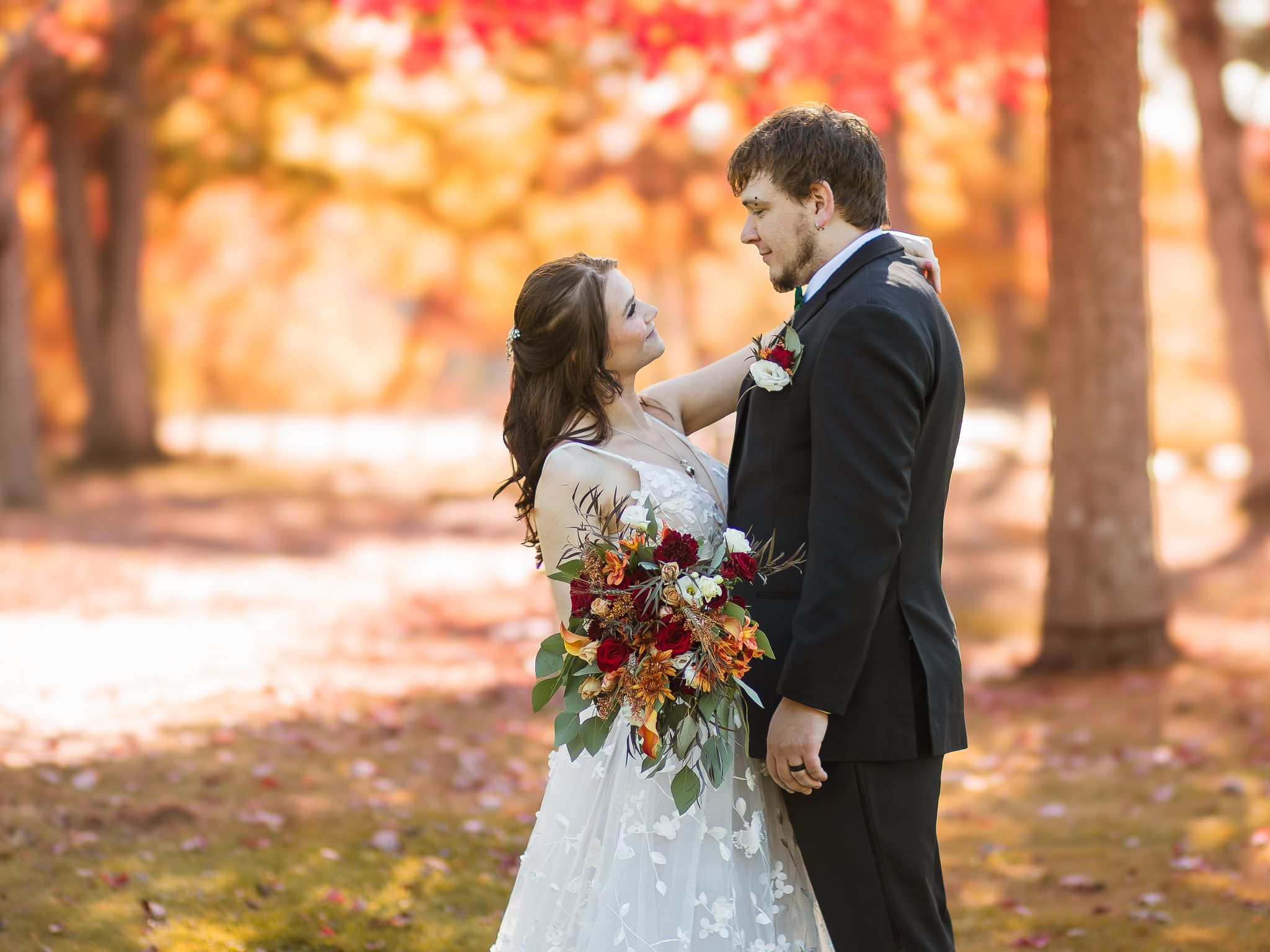A wedding couple stands in a romantic autumn forest setting with vibrant orange foliage and a beautiful fall bouquet.