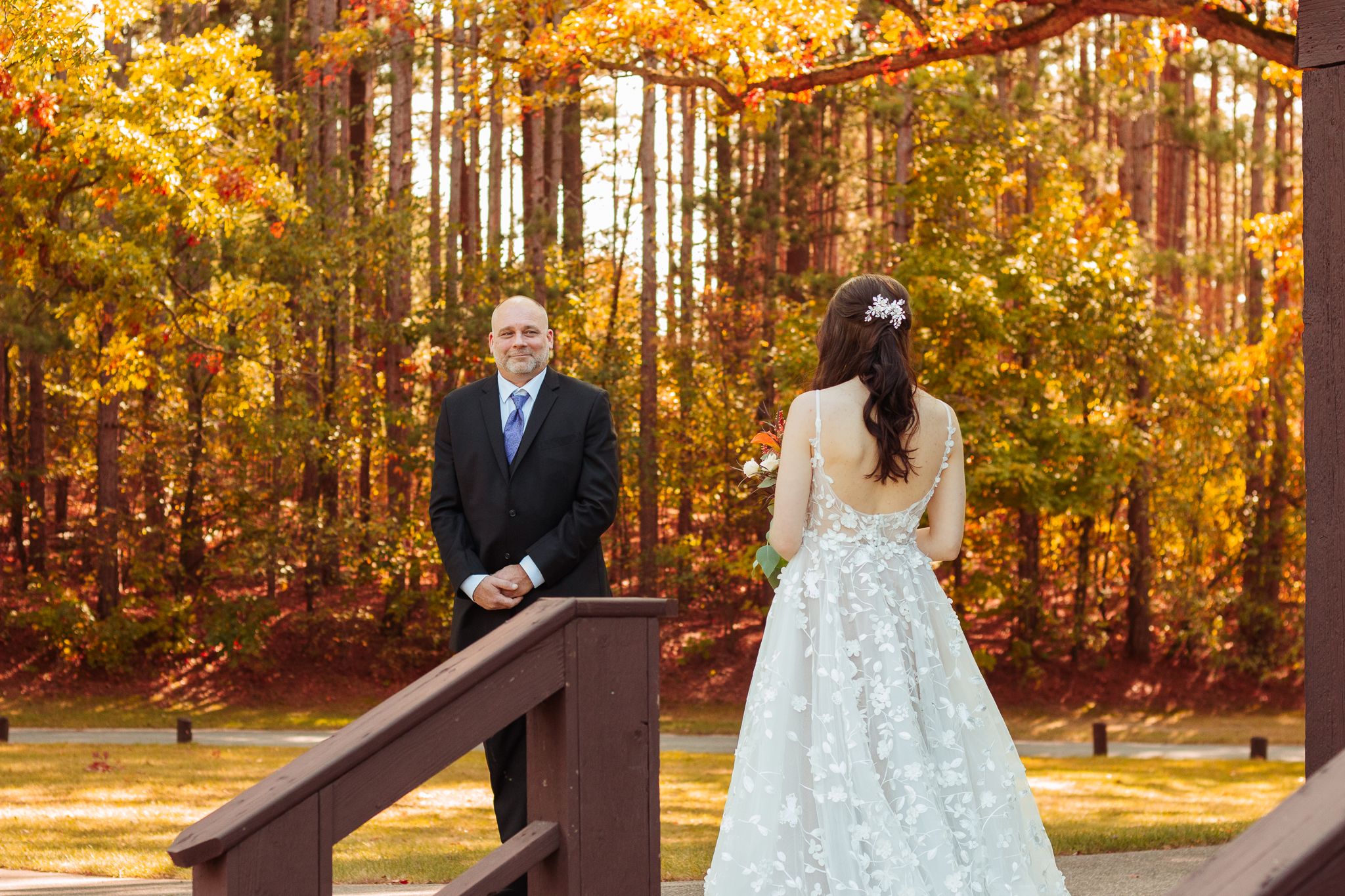 A couple shares a first look moment on a wooden bridge surrounded by golden autumn foliage in the forest.