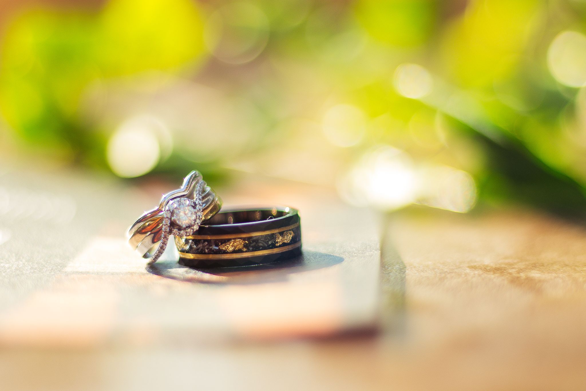 Two gold wedding bands and an engagement ring sparkle on a natural wooden surface with soft green bokeh in background.