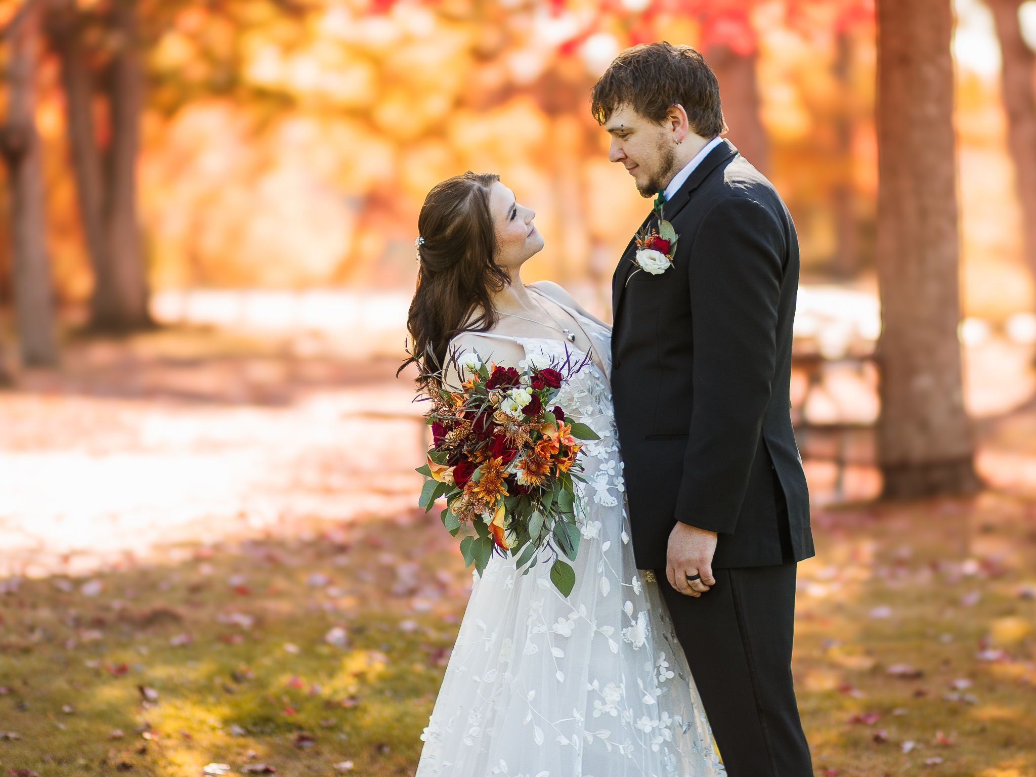 A romantic wedding portrait features a couple standing close together in a forest filled with warm autumn colors.