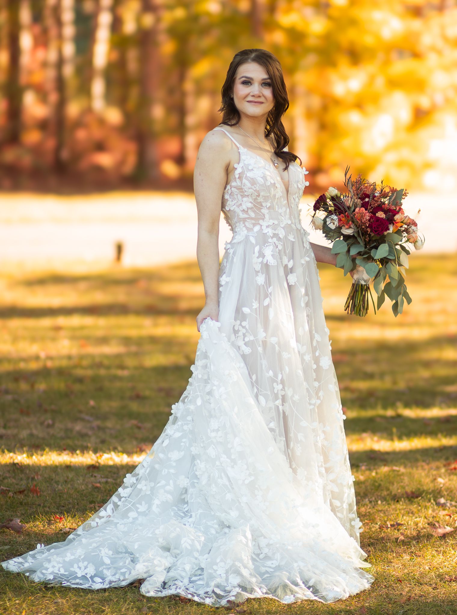 Lace wedding dress with floral appliques and flowing train captured in autumn setting with golden foliage backdrop.