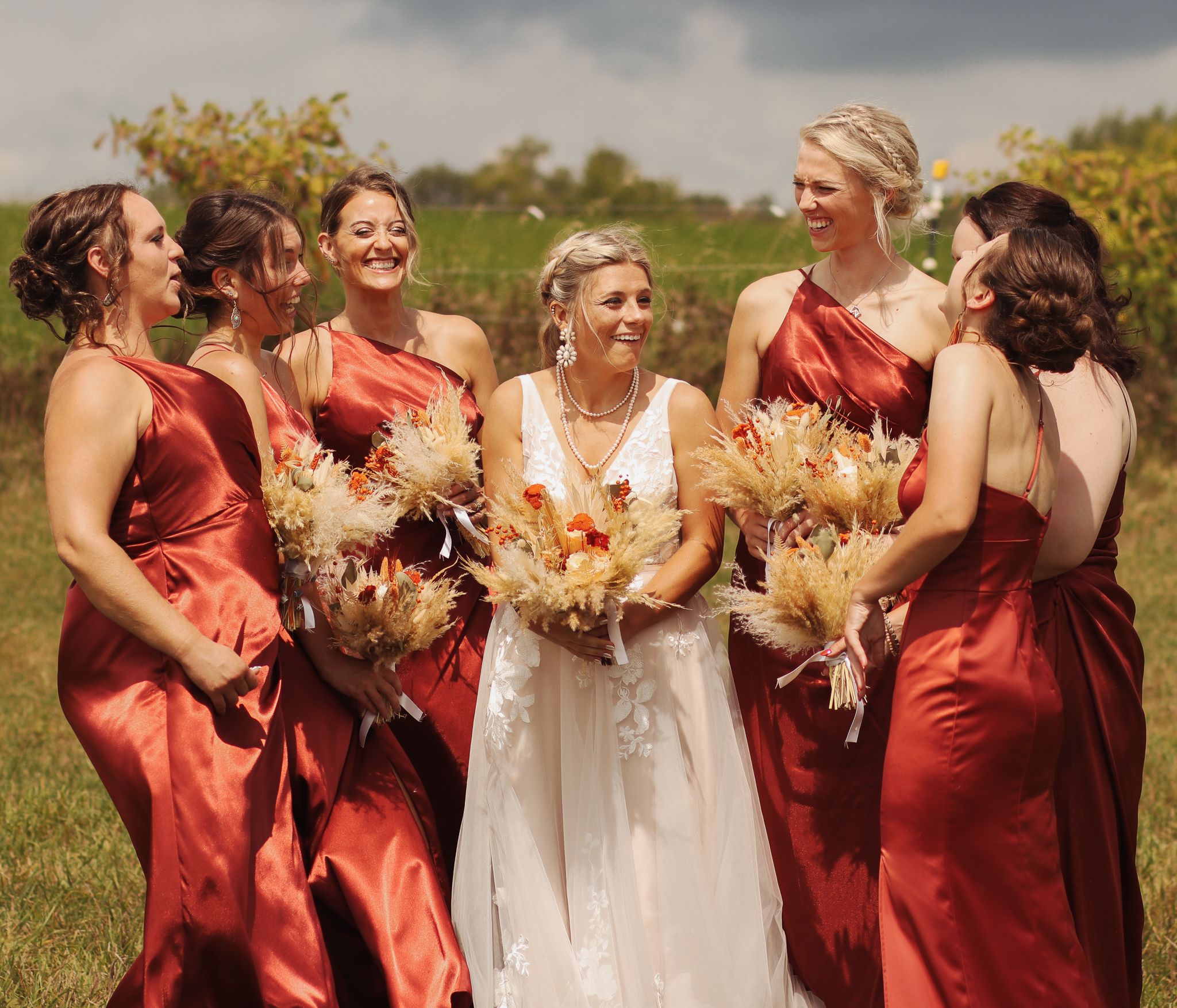 Wedding party in burgundy bridesmaid dresses and white bridal gown sharing joyful laughter in an outdoor field setting.