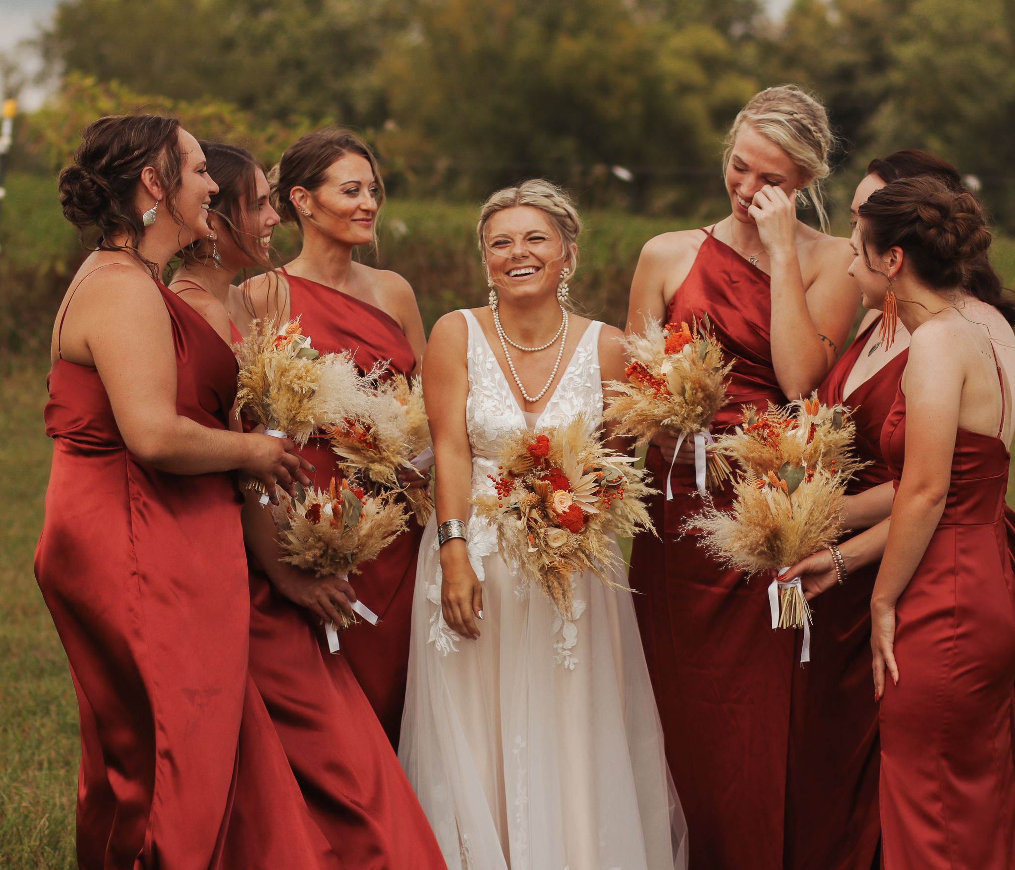 Bridesmaids in red dresses gather around bride with autumn-themed bouquets in outdoor wedding setting.